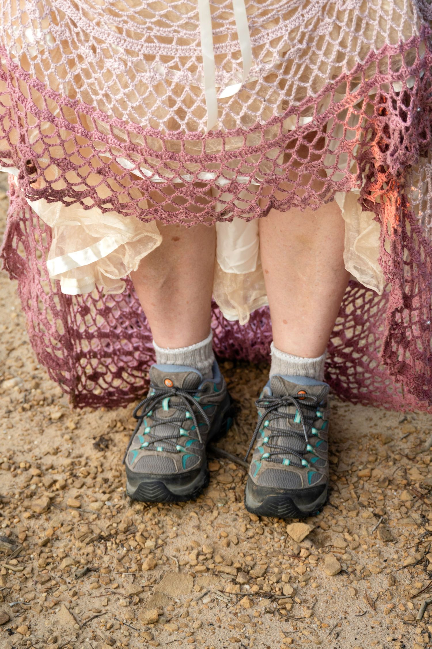 Person wearing hiking shoes and socks under a pink crocheted lace skirt standing on dirt ground.