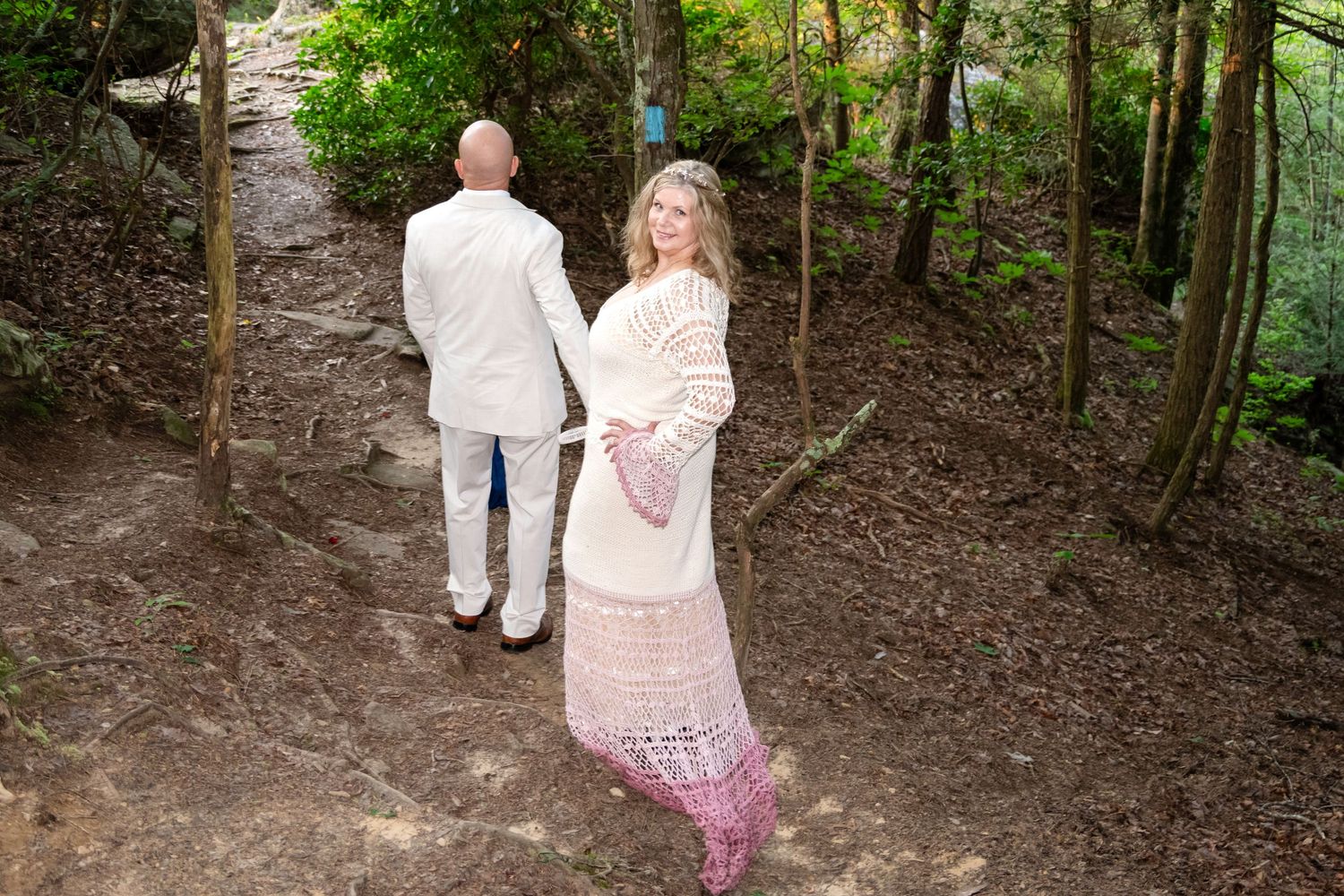 Couple walking on a forest path, woman in pink crochet dress looks back, man in white suit ahead.