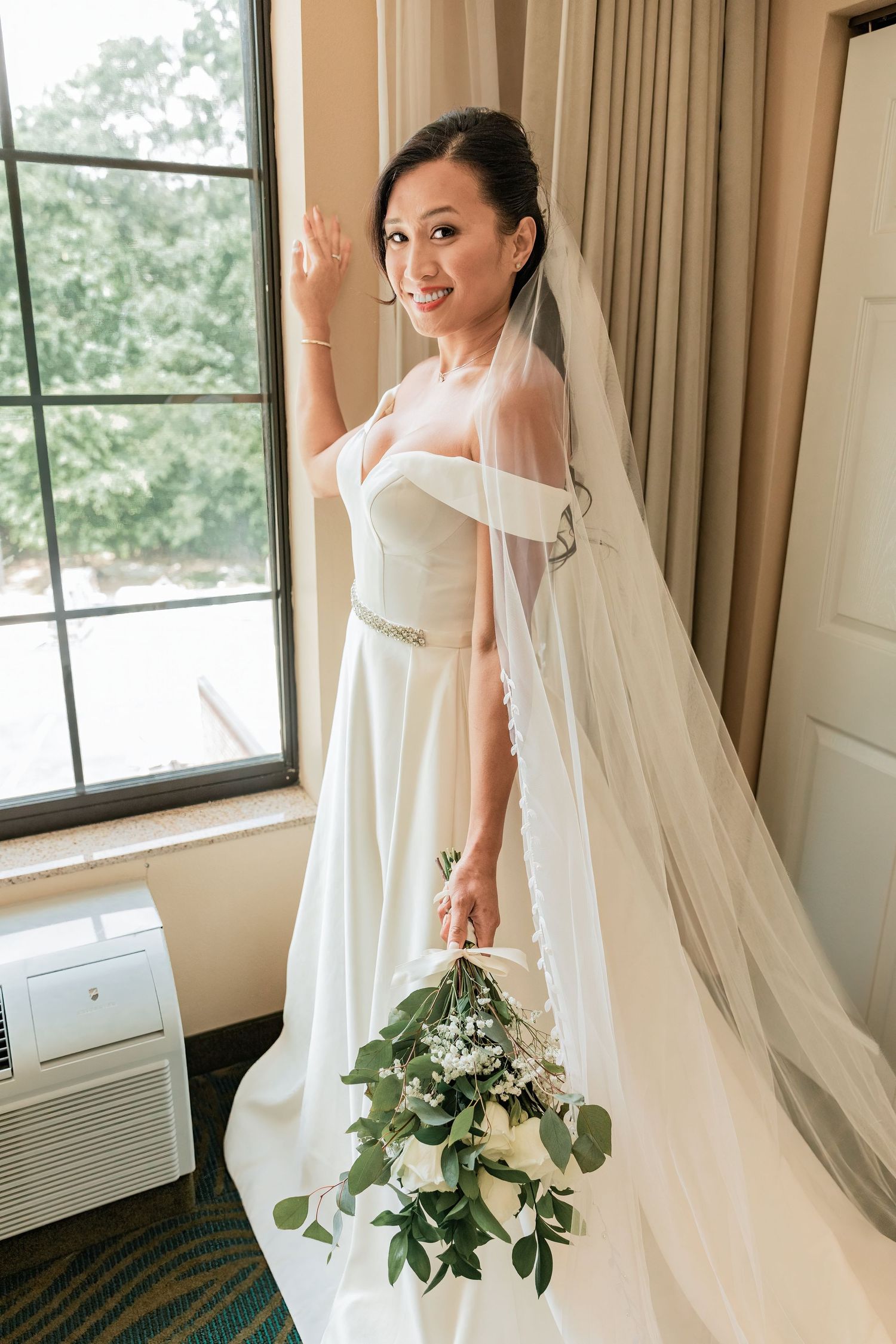 Bride in white off-shoulder gown with long veil holds bouquet of white flowers and greenery, waving near a window.