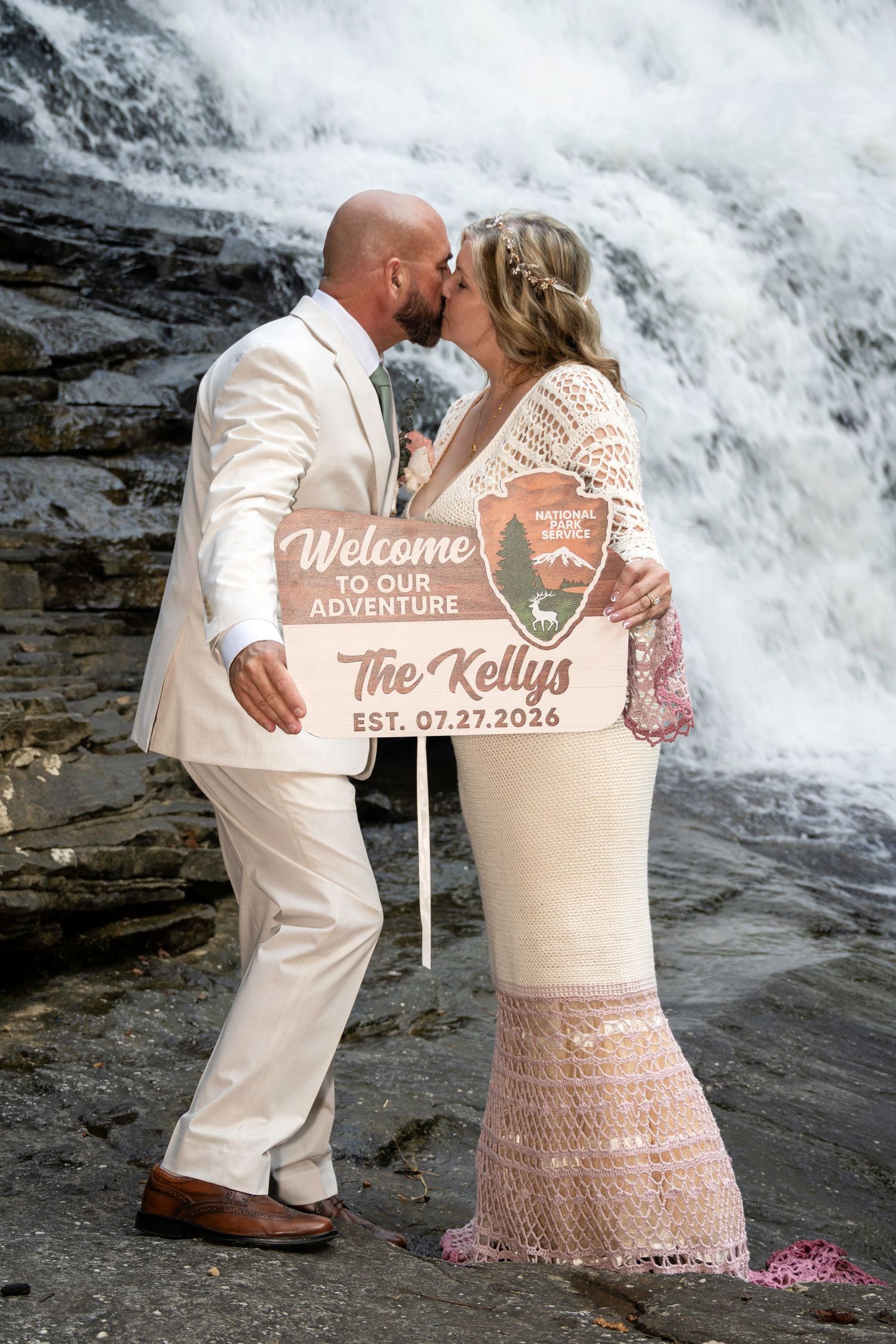Newlyweds kiss in front of a waterfall holding a National Parks wedding sign reading The Kellys Est. 07.27.2026.