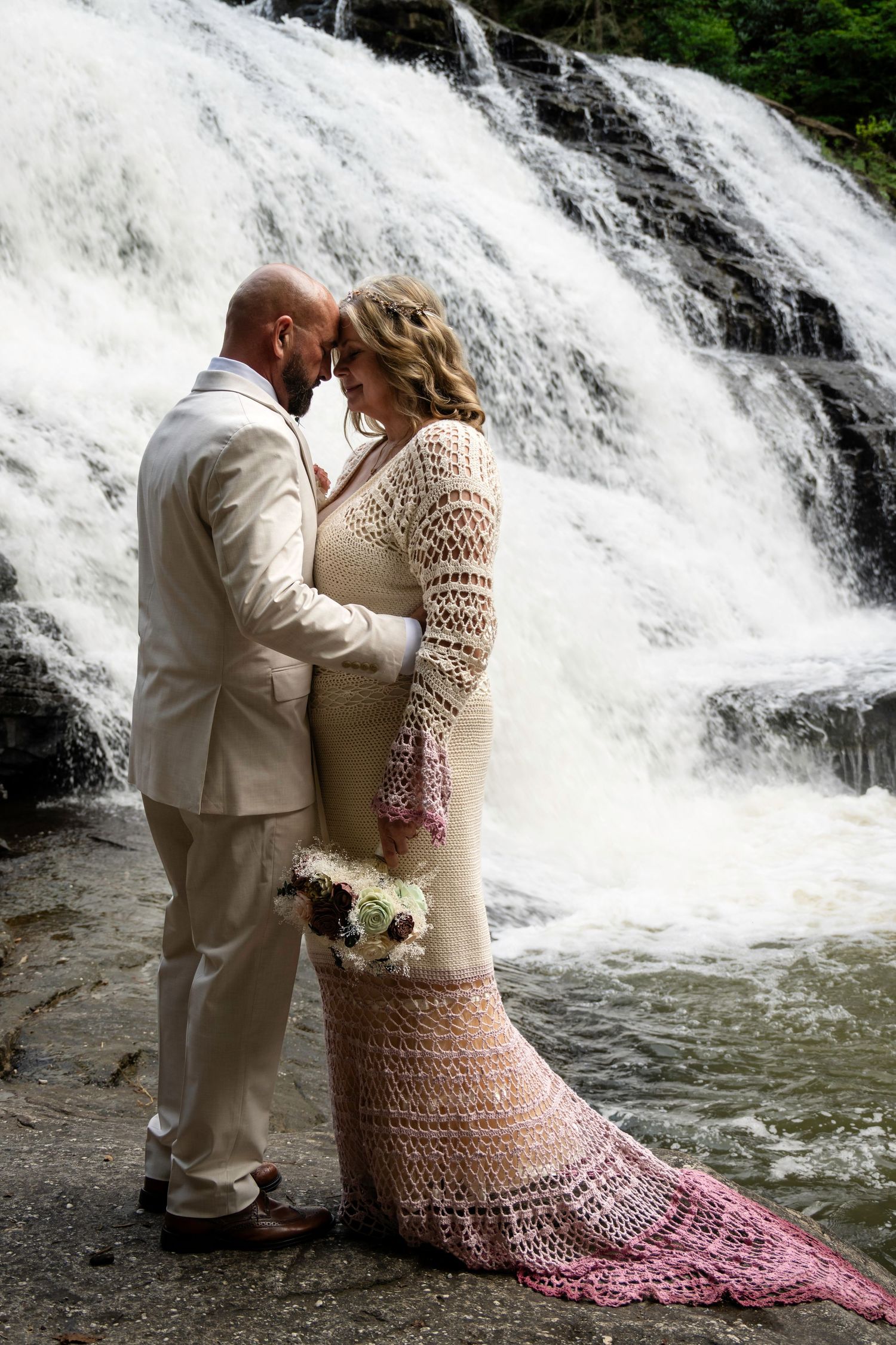 Bride and groom share a romantic moment in front of a cascading waterfall, bride wearing a crochet wedding dress.