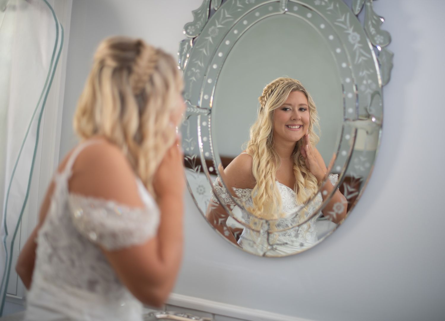 Blonde bride in lace wedding dress smiling at reflection in ornate oval mirror.