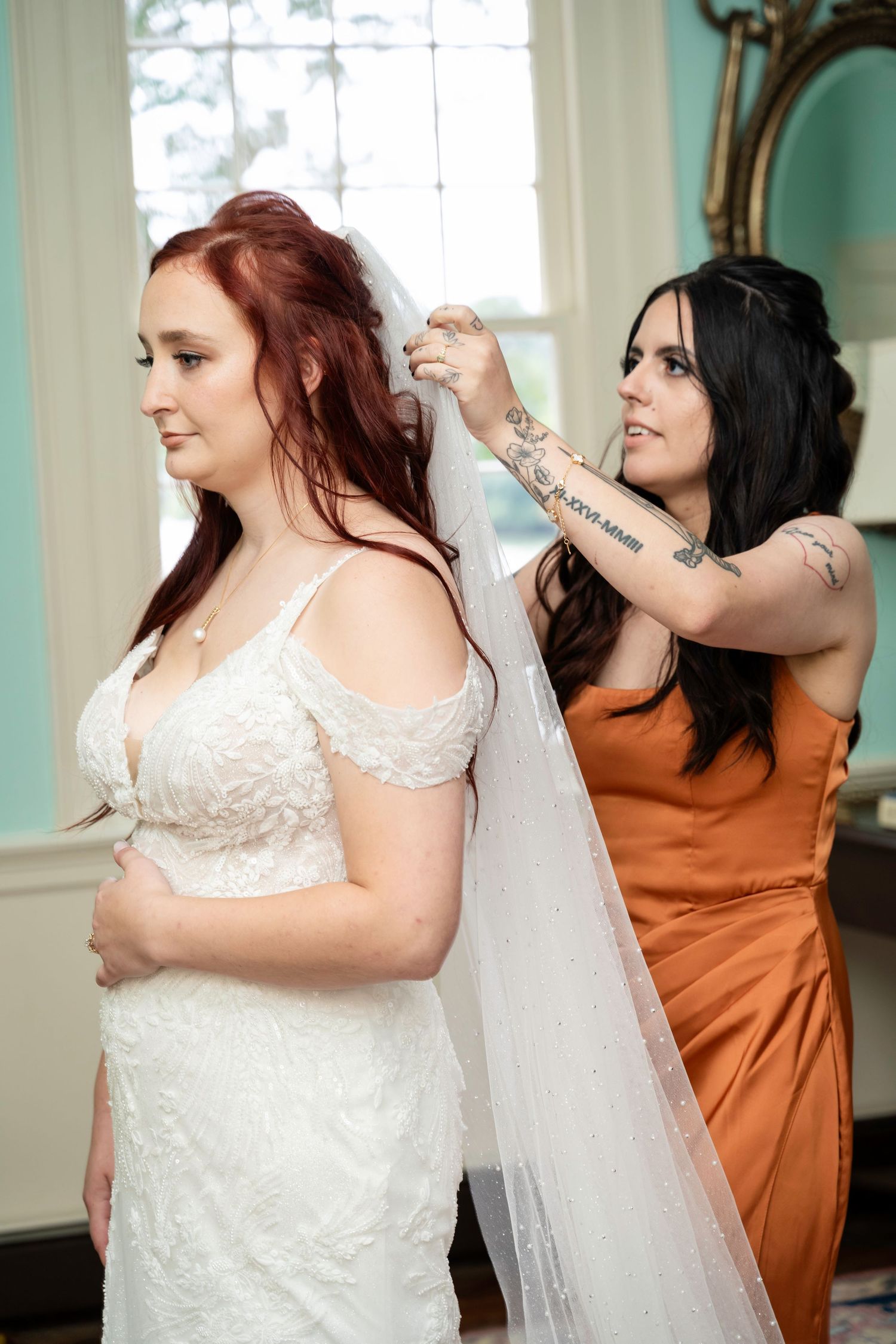 A tattooed bridesmaid in an orange dress attaches a veil to a red-haired bride in a white lace gown.