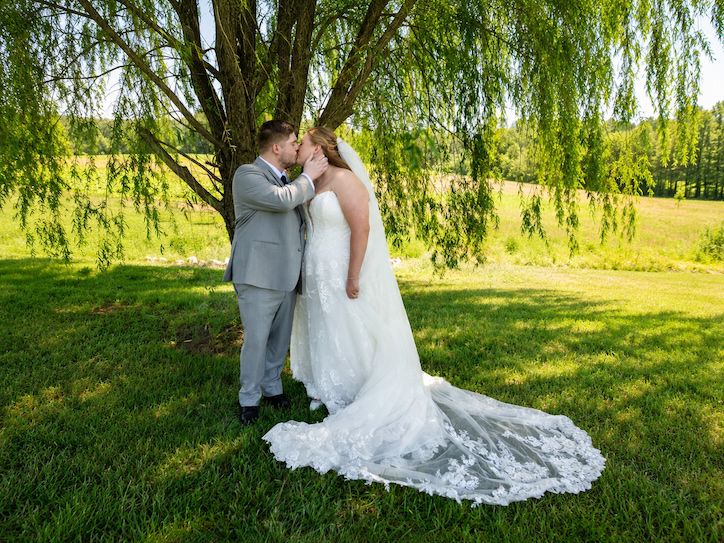 Bride and groom share a romantic kiss under a weeping willow tree on lush green grass on their wedding day.