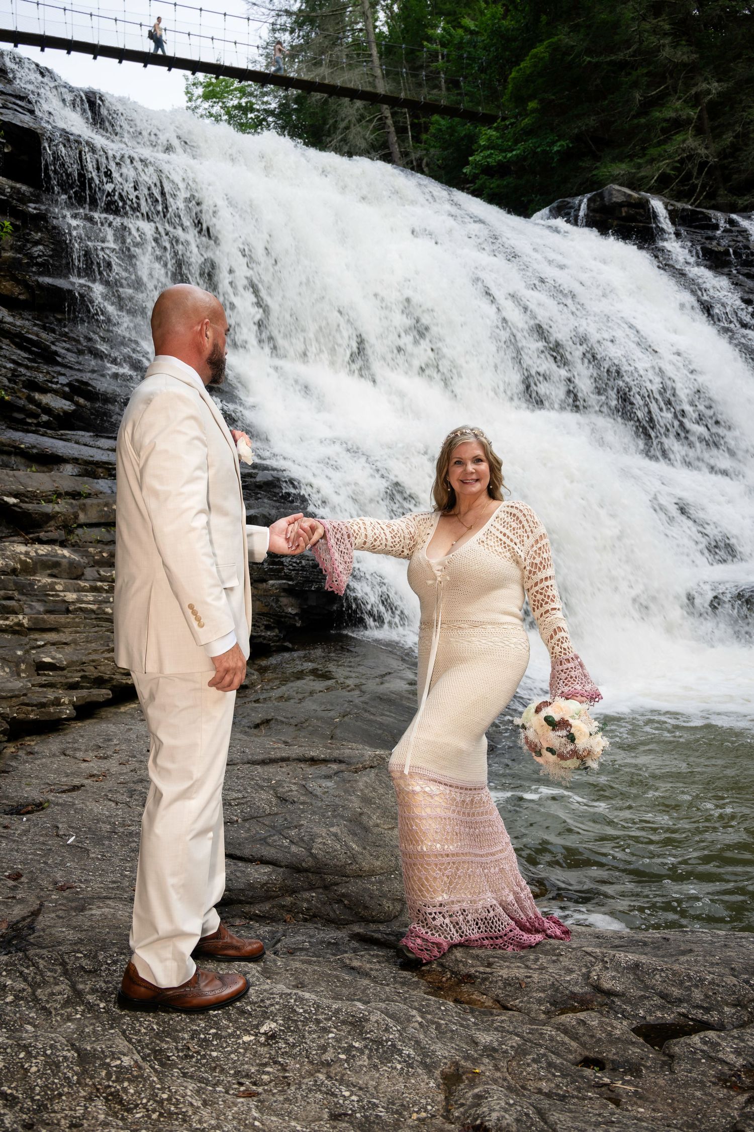 Bride and groom hold hands in front of a stunning waterfall, bride wearing a crochet gown holding a bouquet.