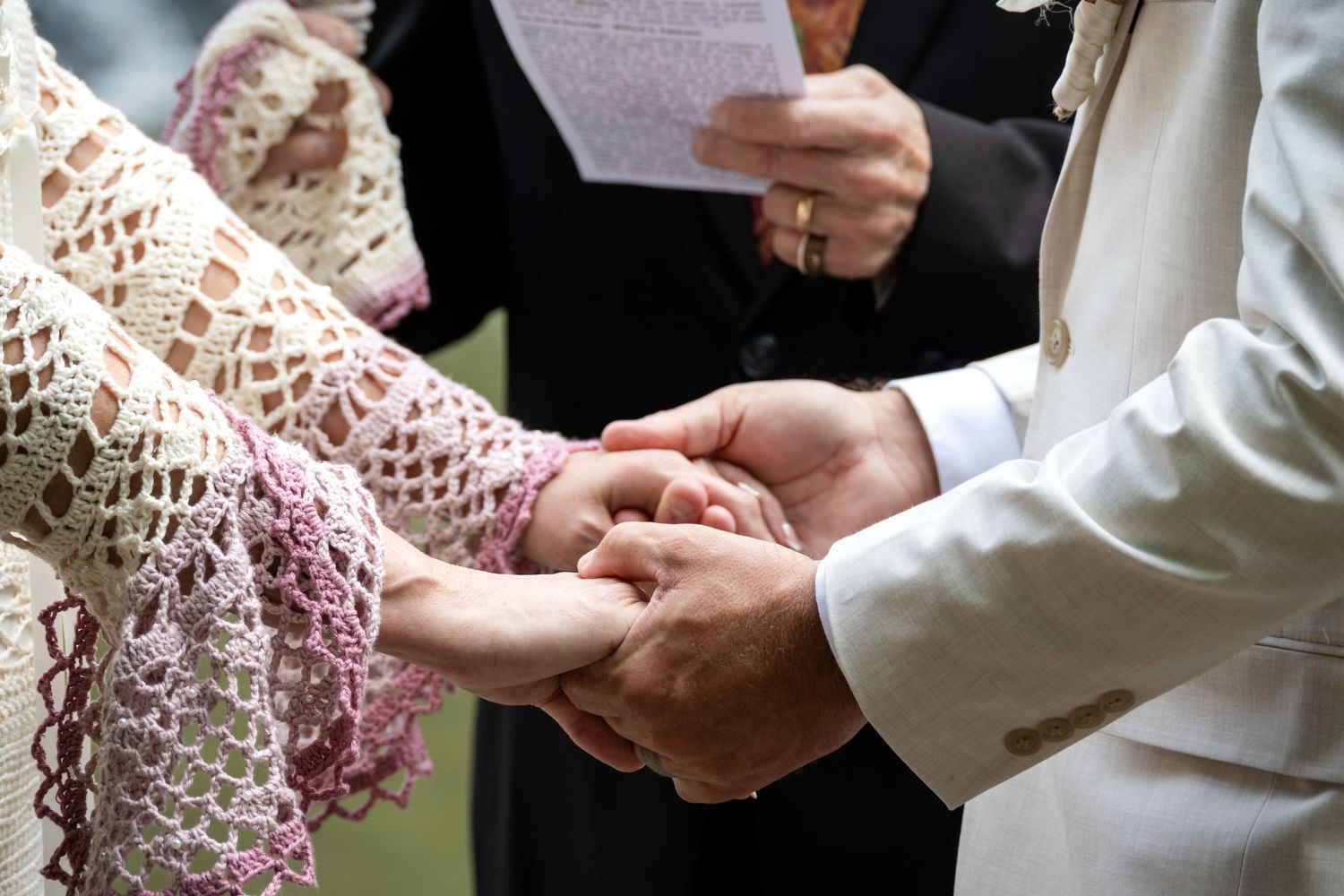 Bride and groom exchanging rings during an outdoor wedding ceremony, officiant holding papers in background.