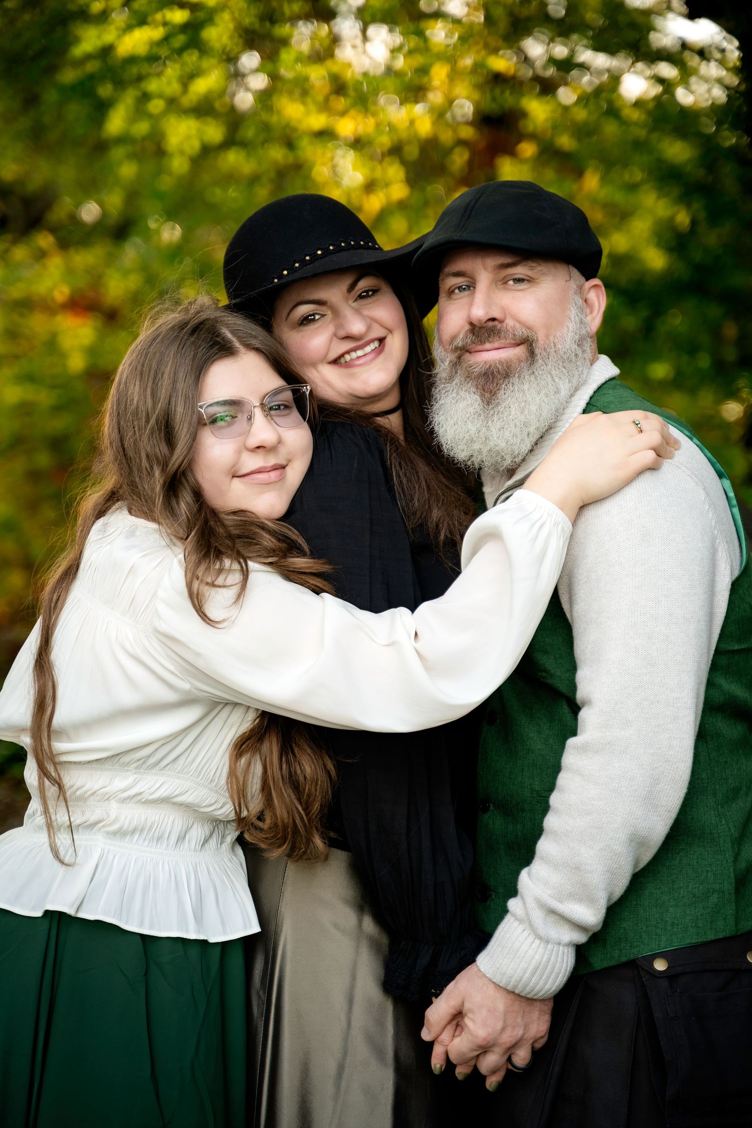 A smiling family of three poses outdoors, with a bearded man, a woman in a black hat, and a teenage girl with long hair.