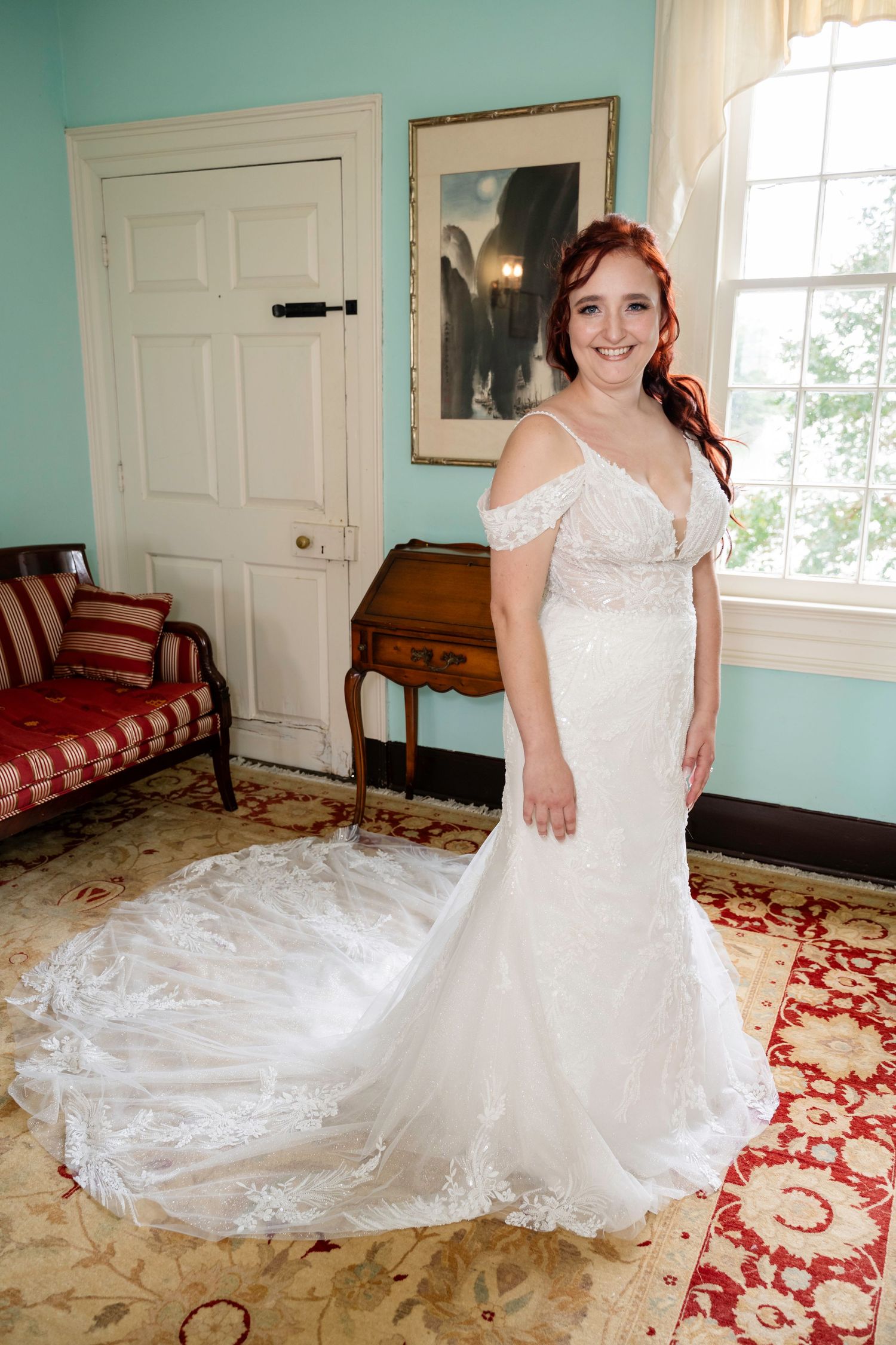 Radiant red-haired bride in off-shoulder lace mermaid wedding gown smiling in vintage room.