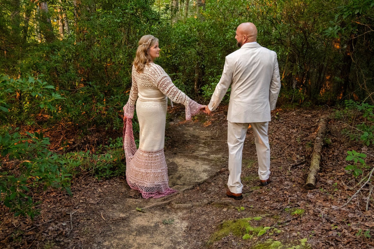 A couple holds hands on a forest path, the woman in a lace gown and the man in a tan suit, viewed from behind.