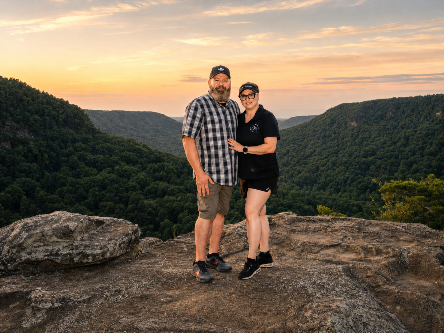 Couple posing on rocky overlook at sunset with lush green valley and colorful sky in background.