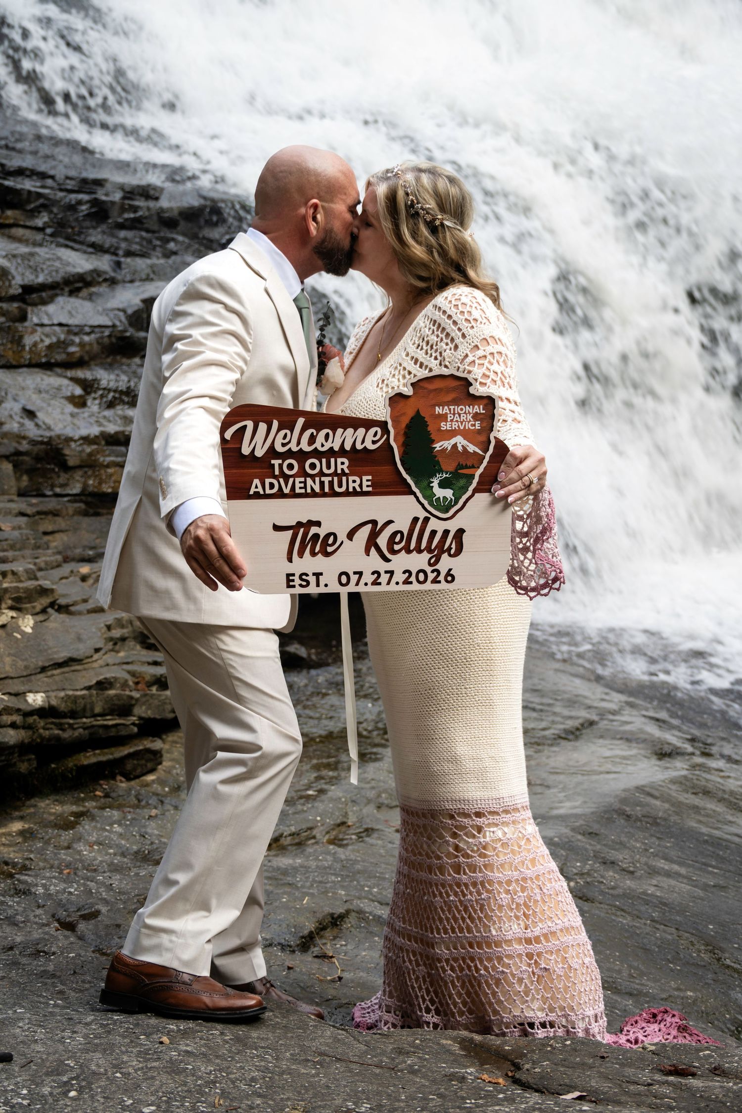 Newlyweds the Kellys kiss by a waterfall holding a National Park Service welcome sign, est. 07.27.2026.
