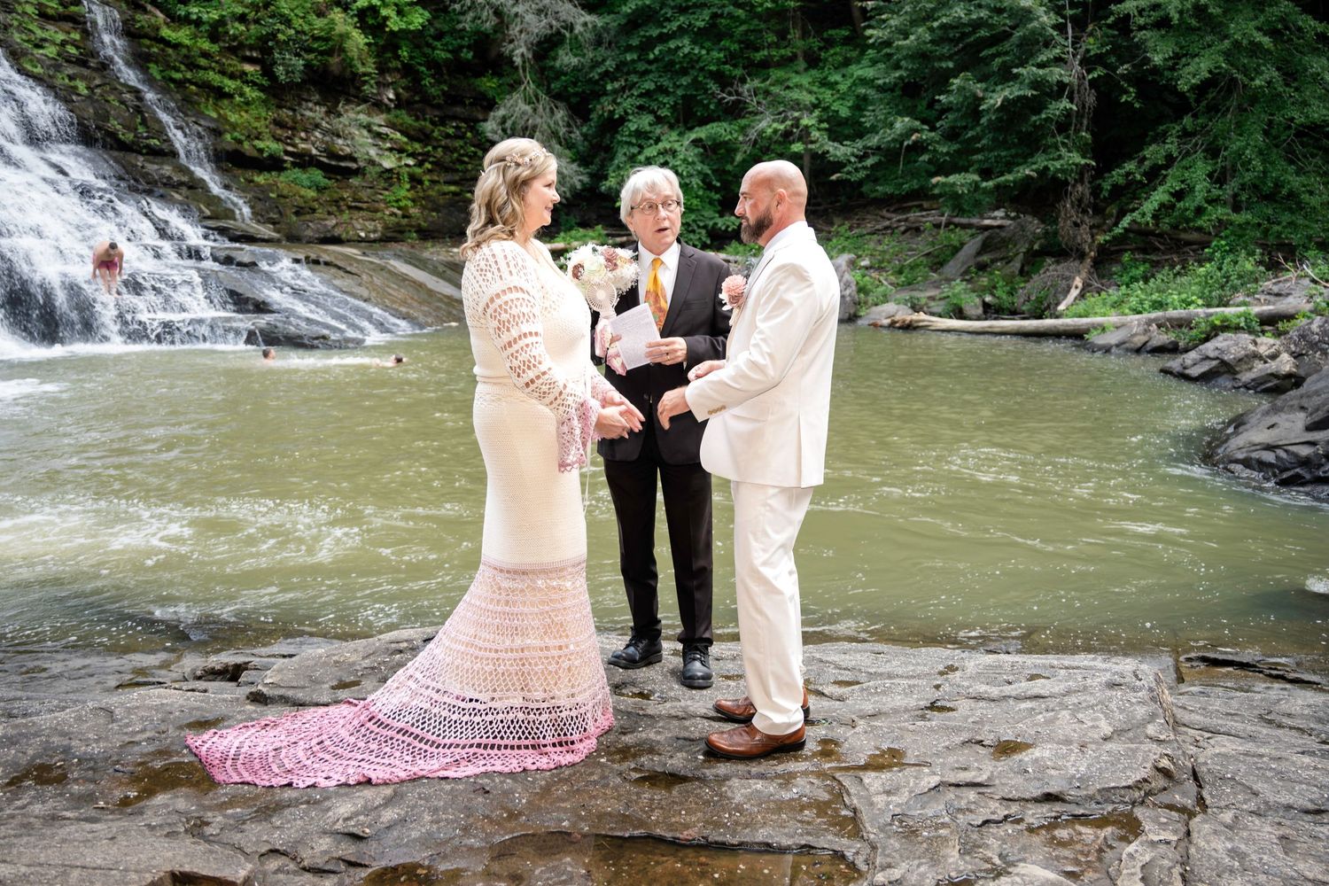 Couple exchanges vows beside a waterfall on rocky terrain, officiated by a minister in a lush green forest setting.
