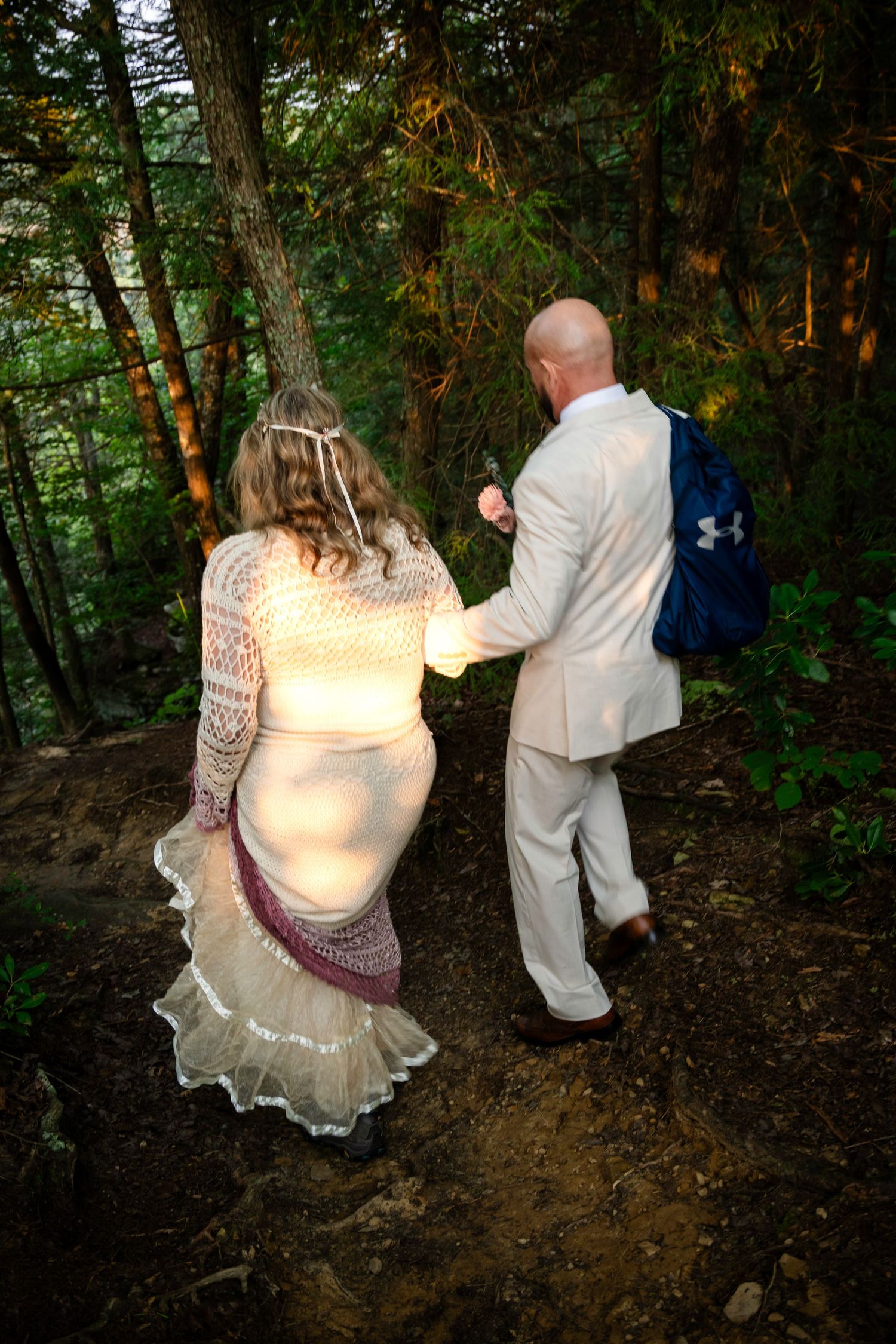 A bride and groom walk hand-in-hand through a lush forest, she in a lace dress, he in a cream suit with a blue backpack.