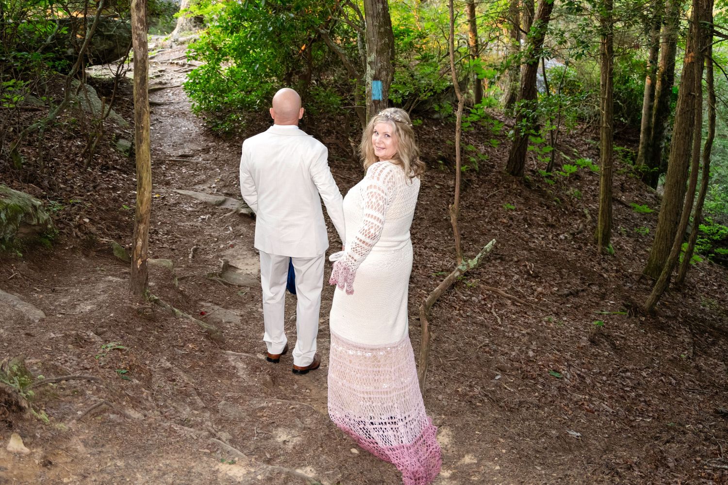 Couple in white wedding attire holding hands on a forest path, woman in lace gown looking back at camera.