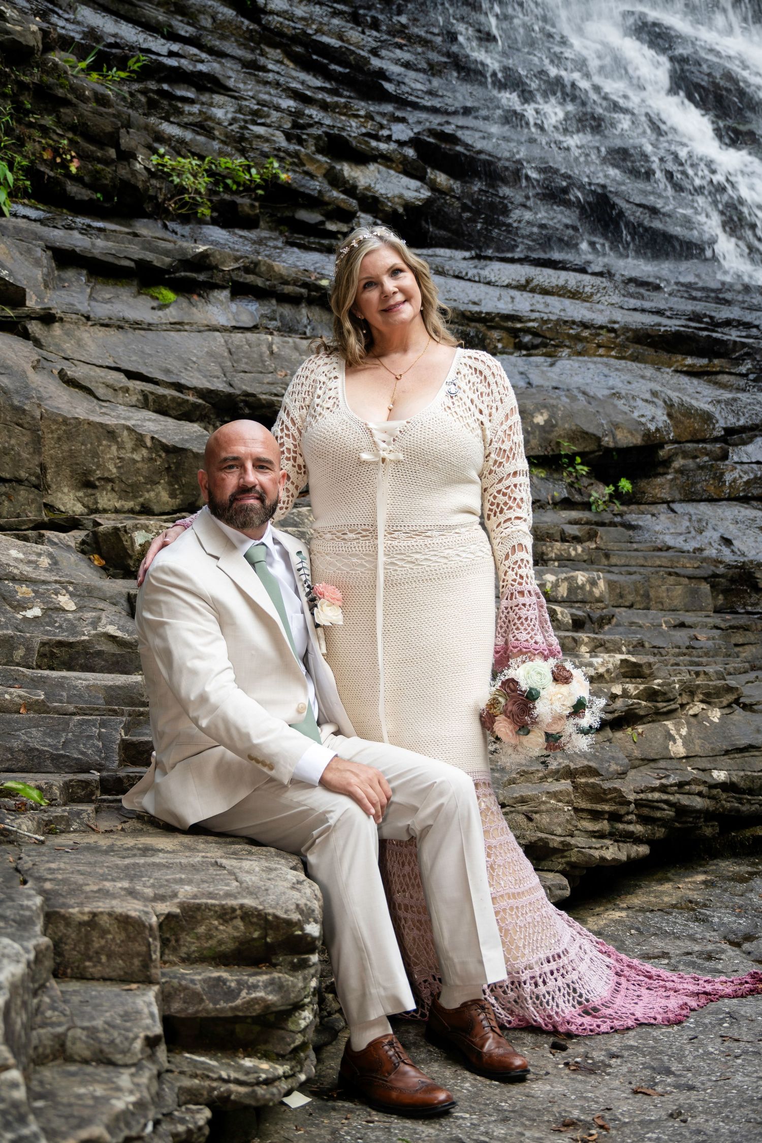 Bride in lace gown and groom in tan suit pose on stone steps before a waterfall for their wedding portrait.