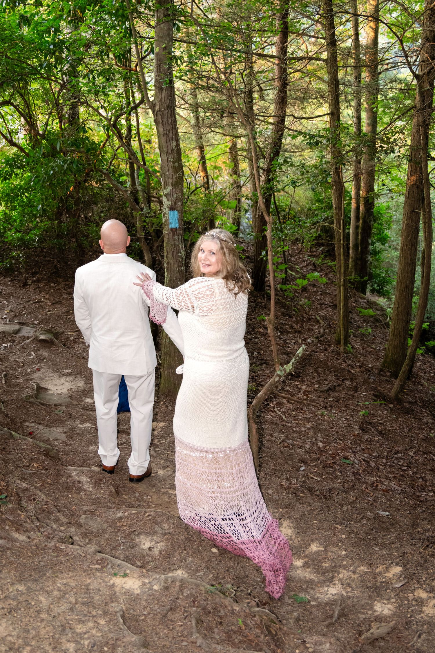A bride in a white lace dress and pink shawl walks with a groom in white suit along a forest trail.