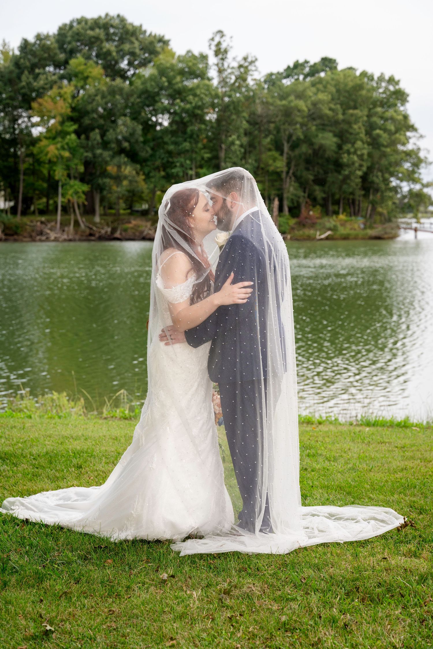 Couple kisses beneath a long dotted veil by the lake, bride in fitted lace gown with train spread on grass.