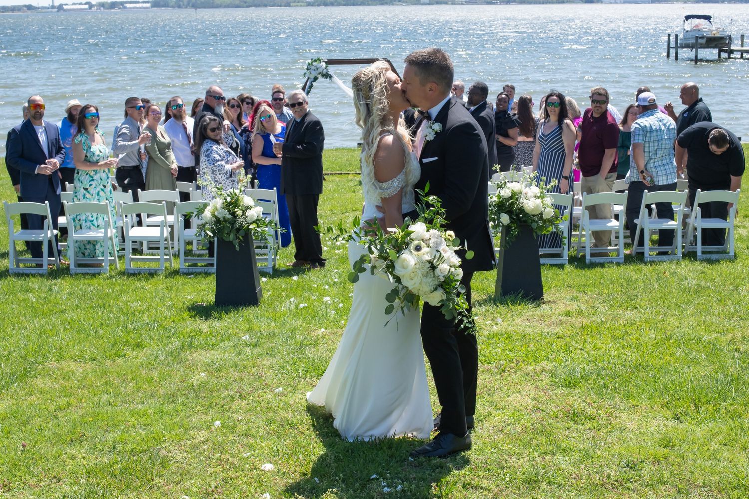 Newlyweds sharing romantic kiss at outdoor lakeside wedding ceremony with guests applauding behind them. - Lusby Wedding Photography MD