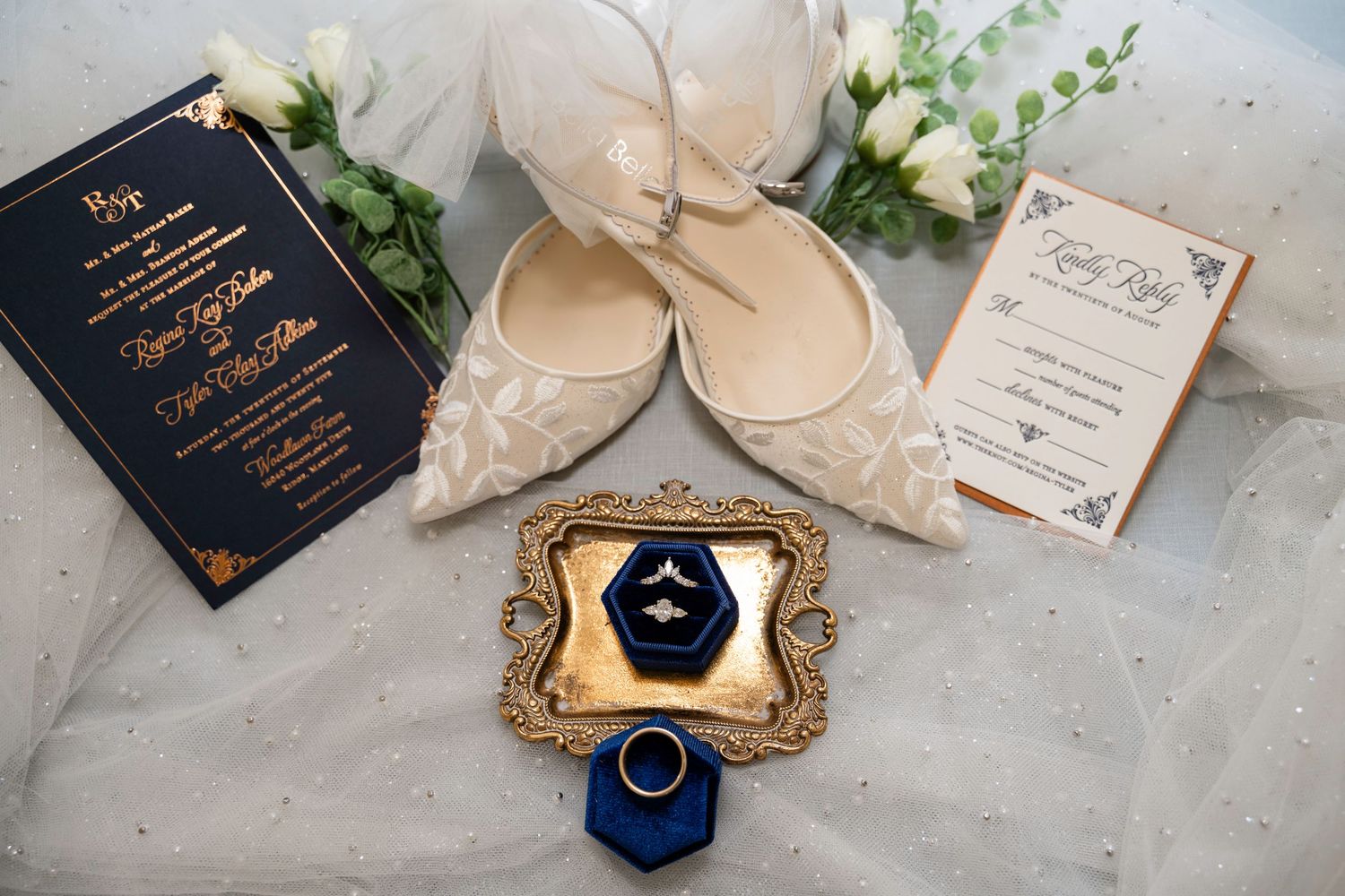 Wedding flat lay featuring ivory heels, navy blue invitations, diamond ring in velvet box, and floral accents.