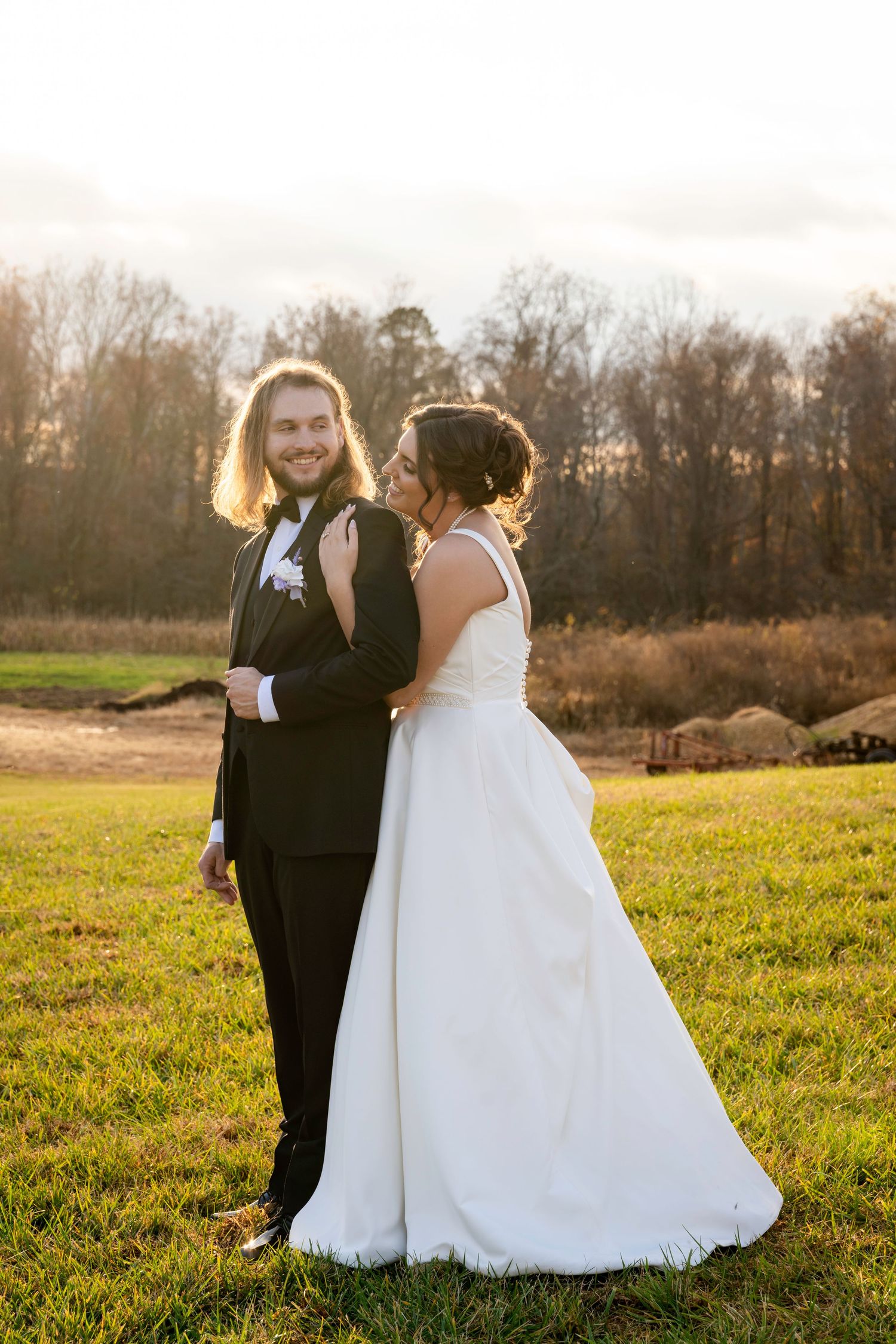 Groom with long hair smiles at camera while bride looks up at him in golden sunset light.