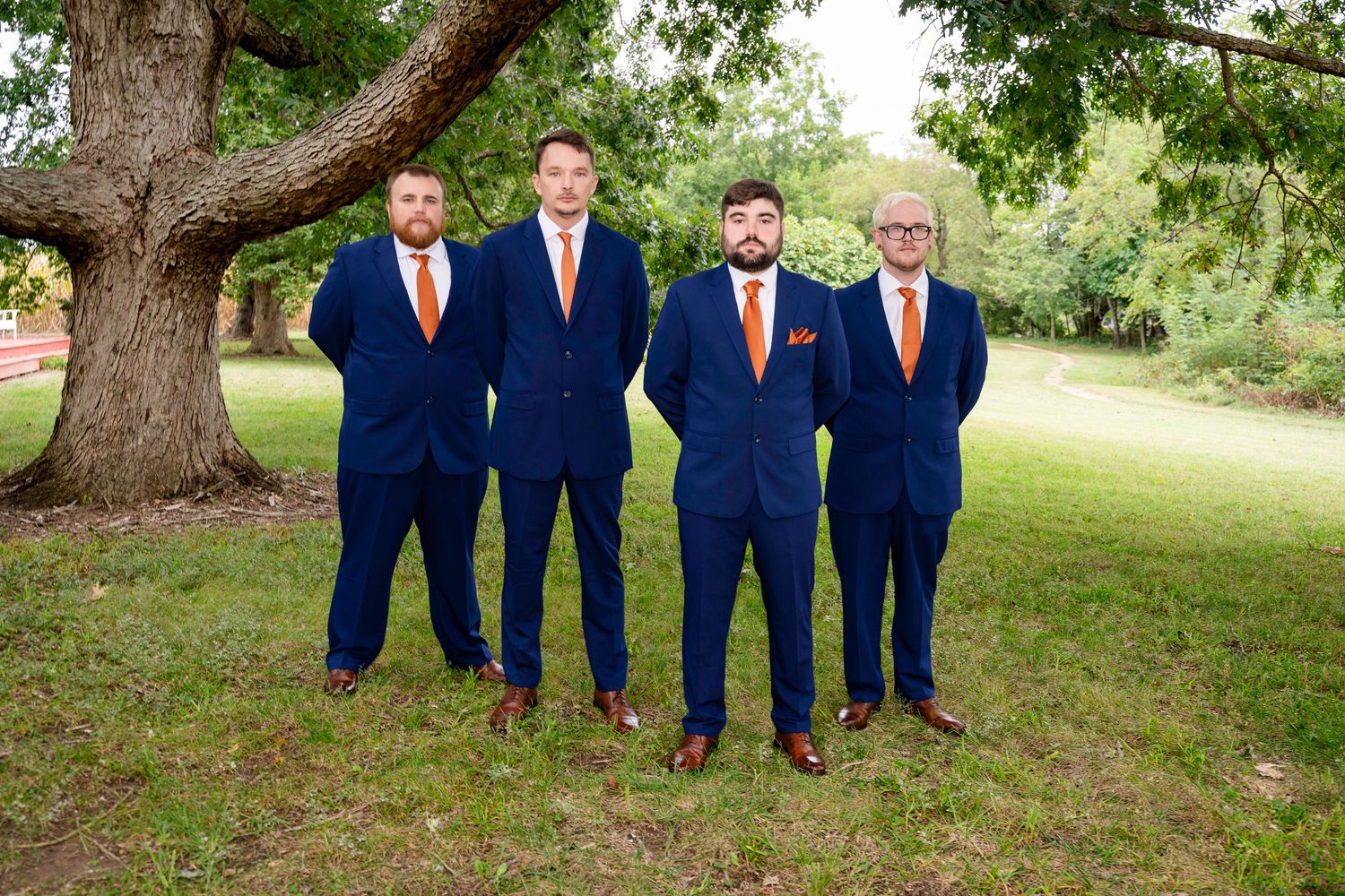 Four groomsmen in matching navy blue suits with orange ties standing together outdoors under a large tree.