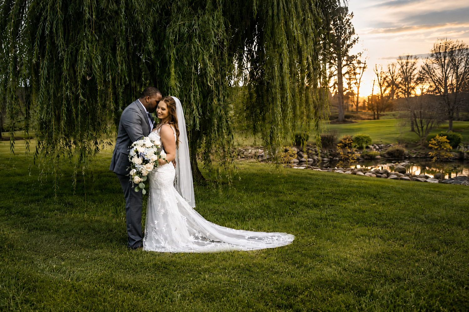 Bride and groom share a romantic sunset moment under a weeping willow tree by a pond on their wedding day.