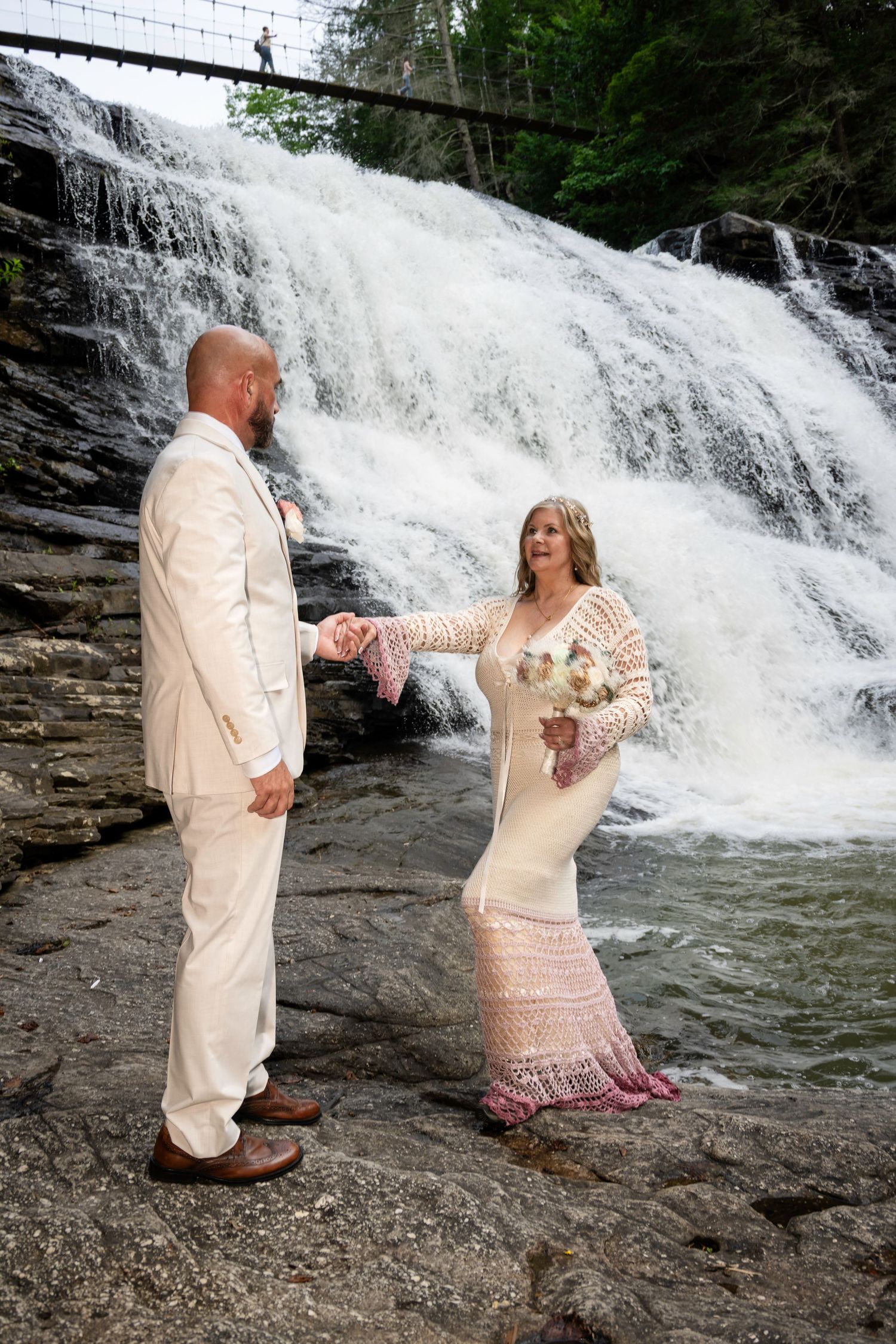 Bride and groom hold hands before a stunning waterfall backdrop, she in a lace gown, he in a cream suit.