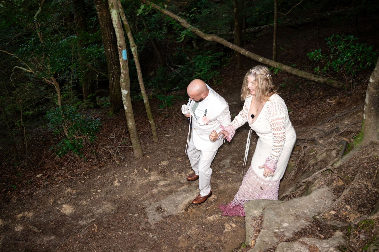 A bald groom in white suit helps his blonde bride in lace dress navigate a forest trail during an outdoor woodland wedding.