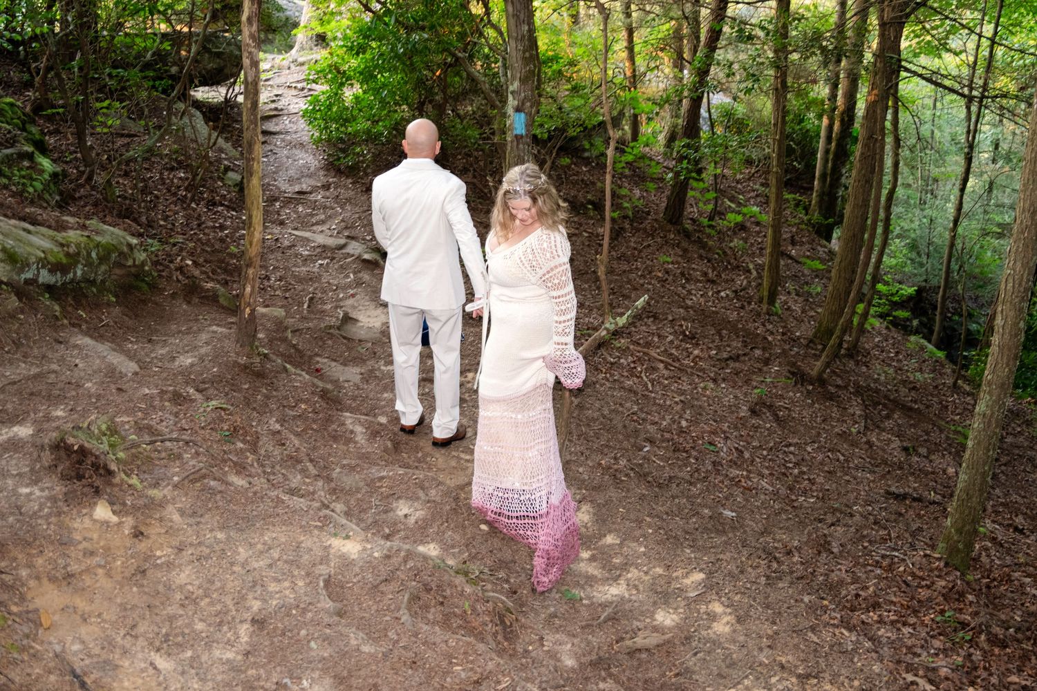 A bride and groom in white wedding attire walk hand-in-hand along a wooded forest trail.