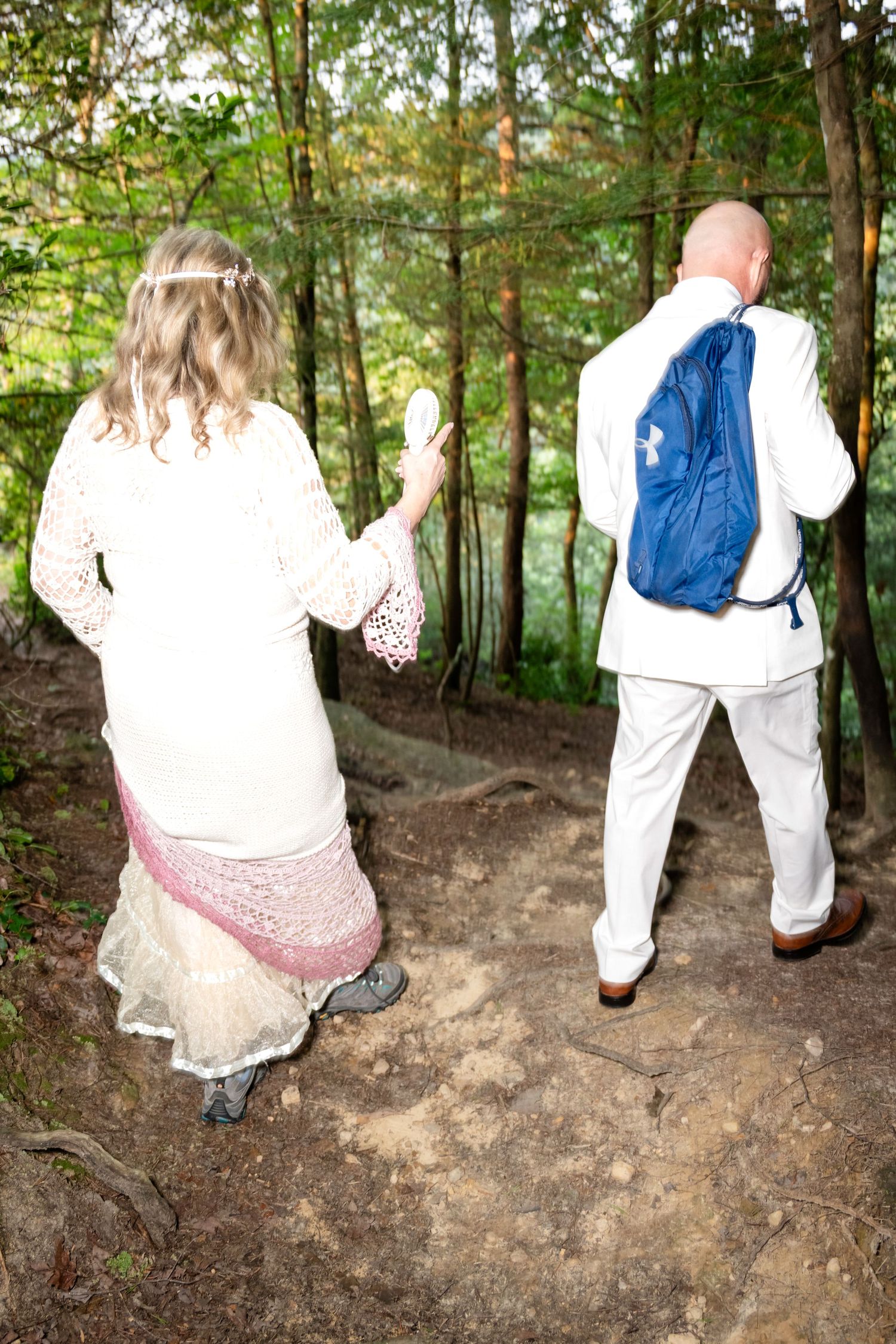 A bride and groom in white wedding attire walk along a forest trail, with the groom carrying a blue backpack.