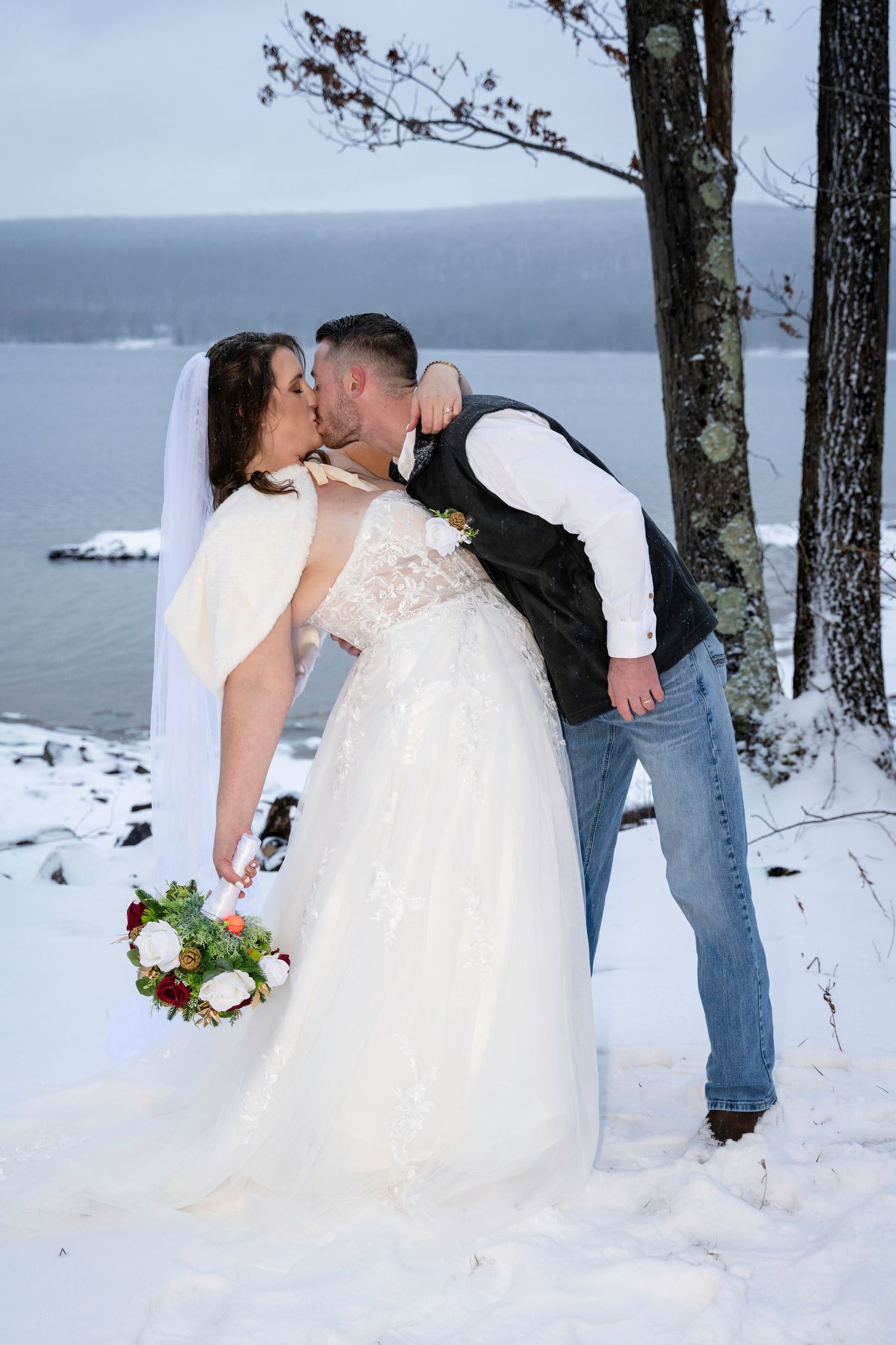 Bride and groom share a kiss in a snowy winter landscape near a lake, bride holding a red and white bouquet.