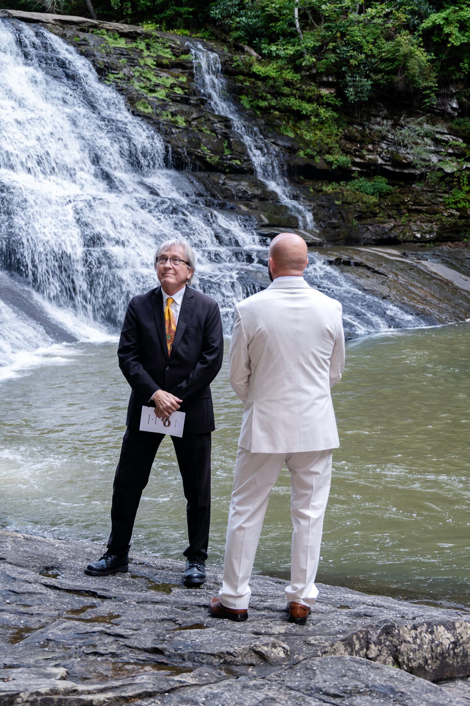 Two men in formal suits stand on rocks before a cascading waterfall during an outdoor ceremony.