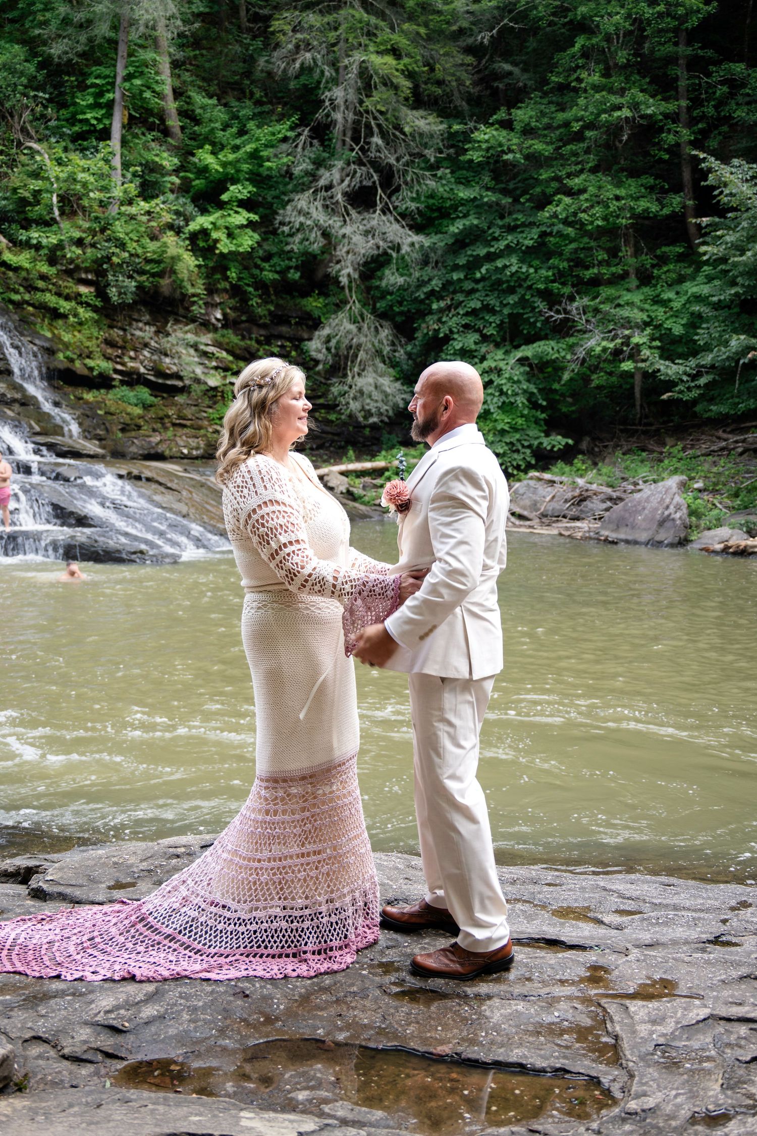 Couple exchanging vows on rocky riverbank with waterfall and lush green forest backdrop, bride in crochet gown.