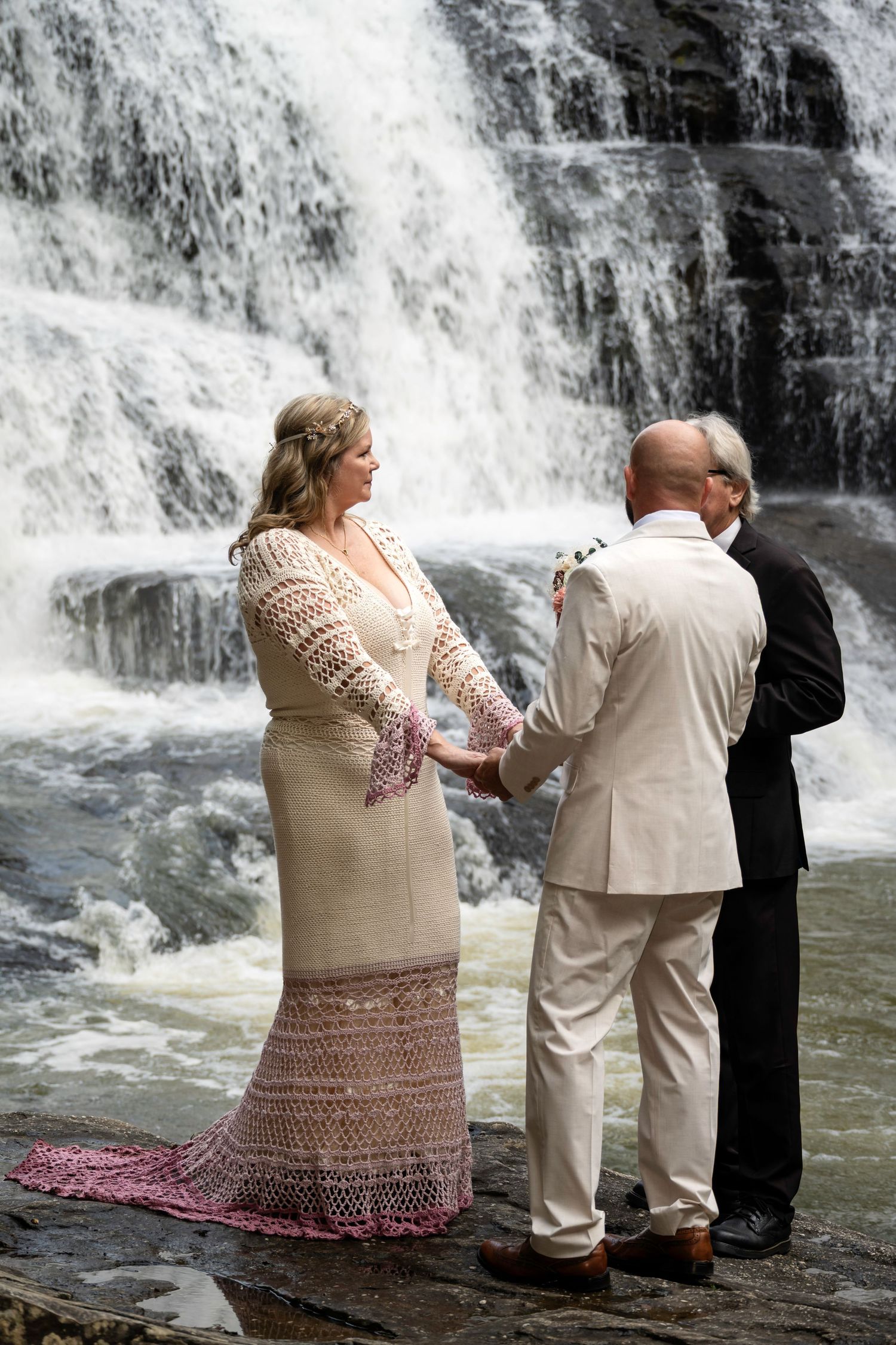 Couple exchanges vows before a stunning waterfall backdrop, bride in lace gown, groom in cream suit, officiant presiding.