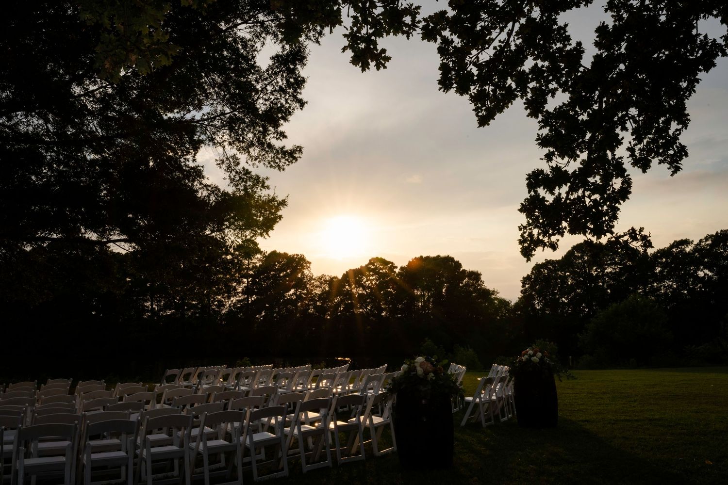 Sunset silhouettes rows of white chairs at an outdoor wedding ceremony framed by sweeping tree branches.