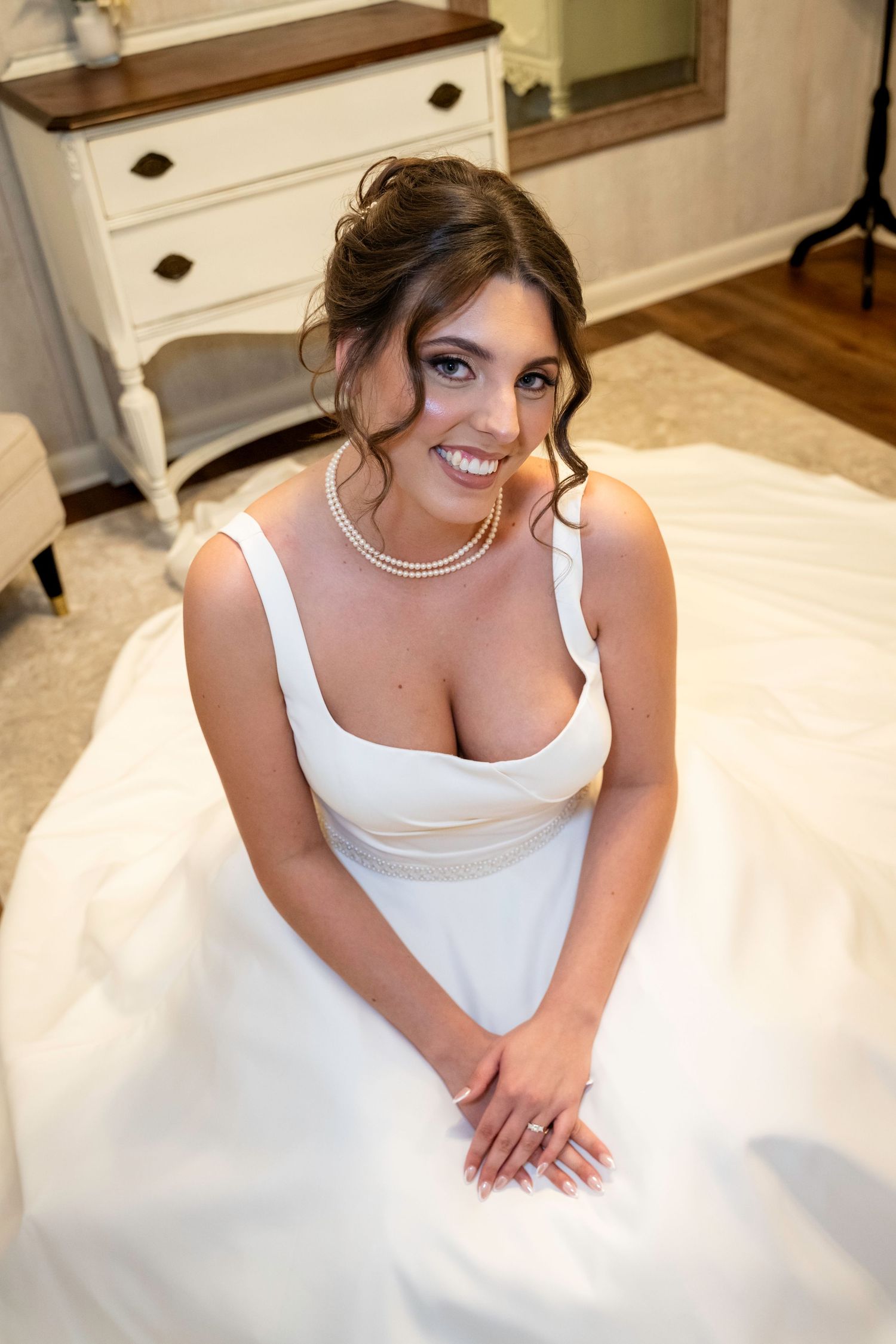 Beautiful bride in elegant white wedding gown with pearl necklace sitting on floor smiling at camera.