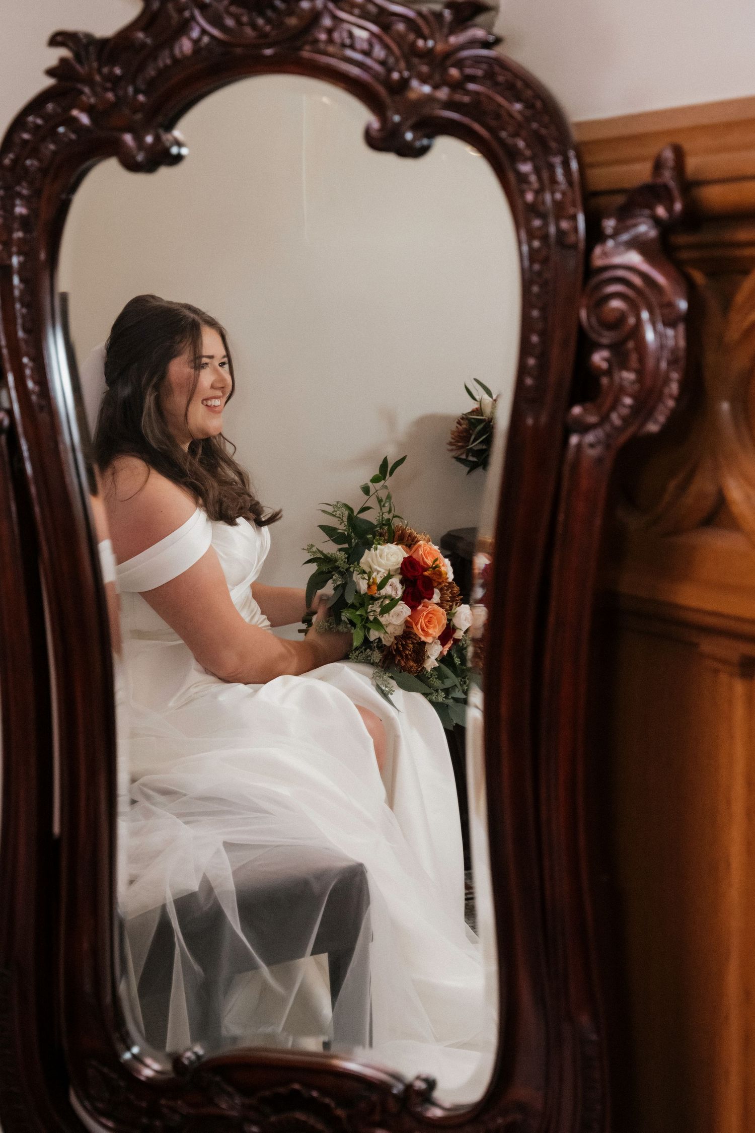 Smiling bride in off-shoulder white gown holding colorful bouquet reflected in ornate antique carved wooden mirror.