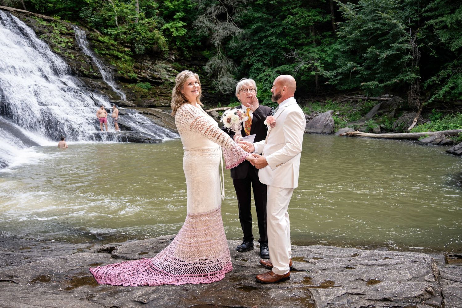 Couple exchanges vows before a stunning waterfall backdrop, bride in pink lace gown, groom in cream suit.