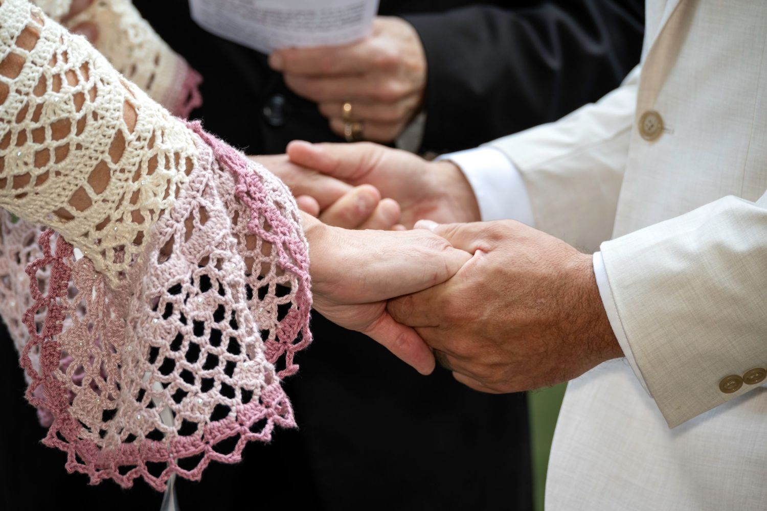 Couple exchanging rings during a wedding ceremony, bride wearing pink lace shawl, groom in white jacket.