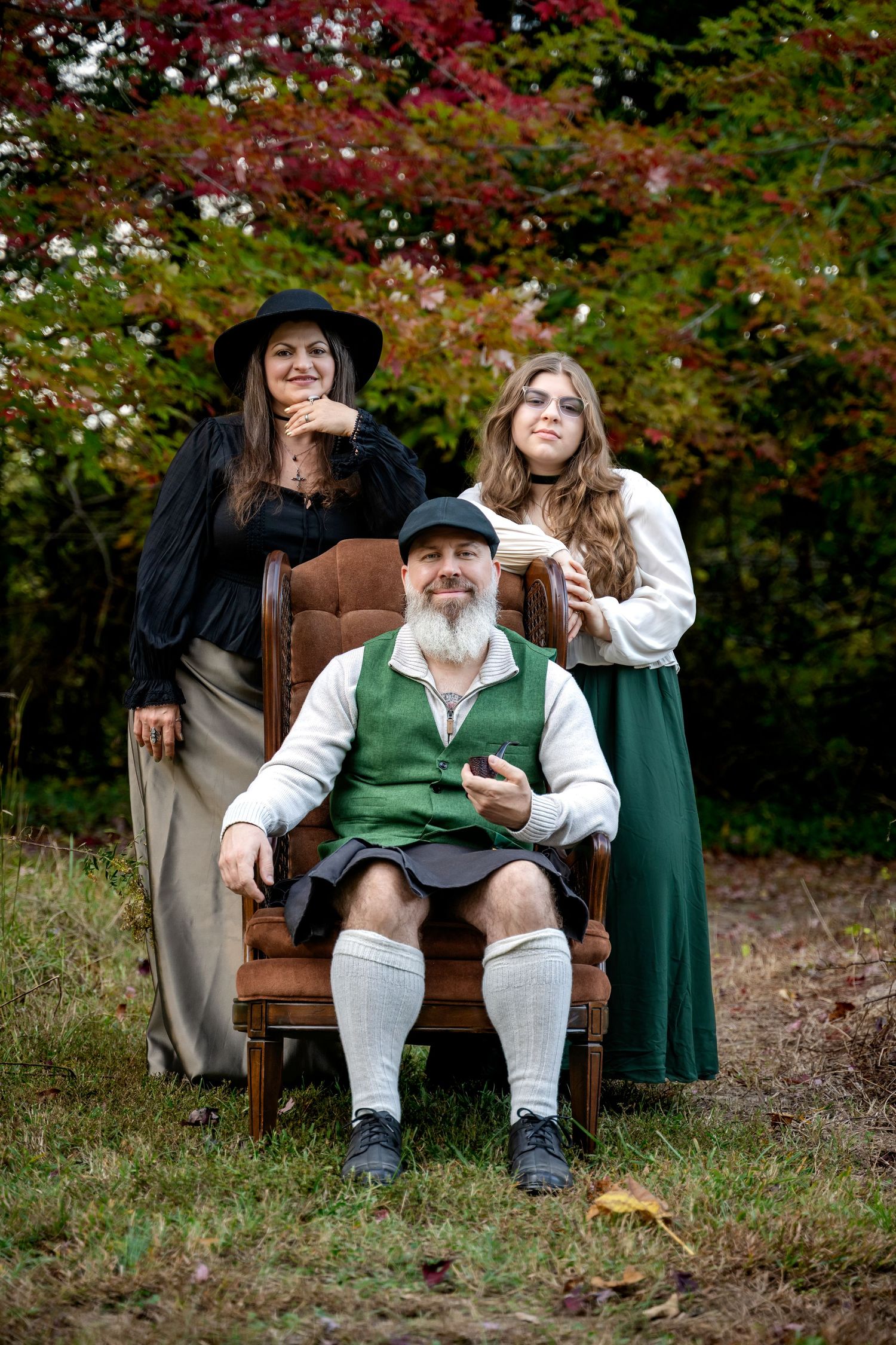 Man in Scottish attire sits in vintage chair outdoors, flanked by two women in period costumes, with autumn trees behind.
