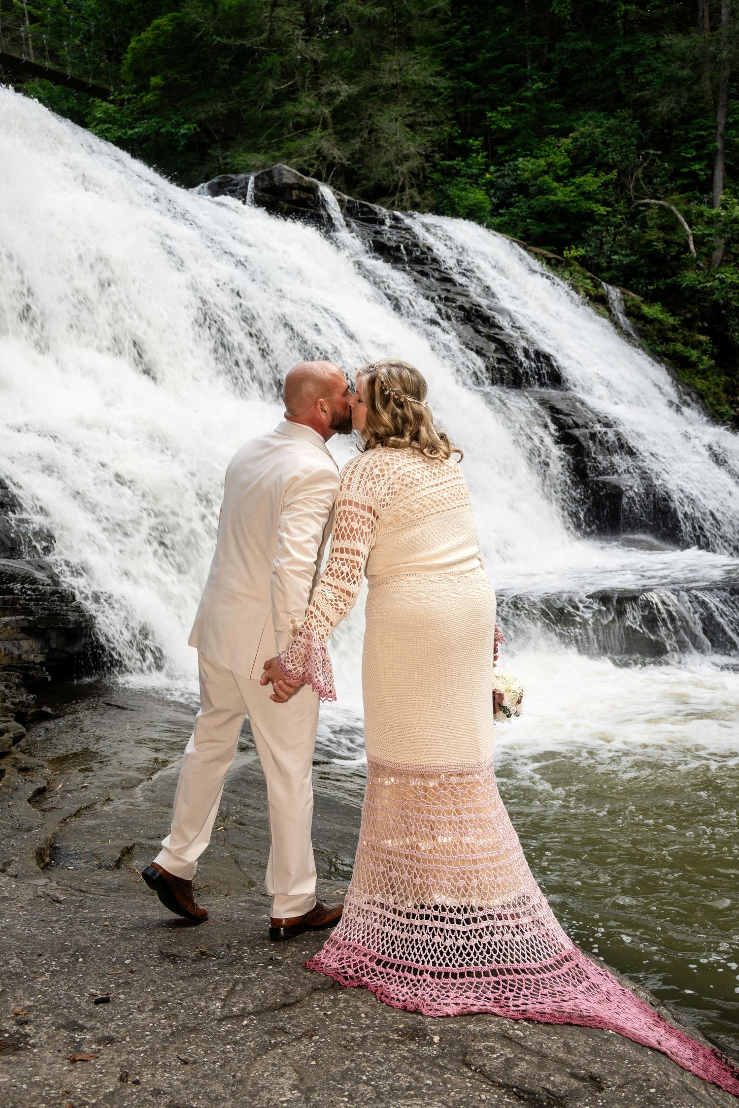 Bride and groom kissing in front of a cascading waterfall, bride wearing a crochet wedding dress with pink hem.