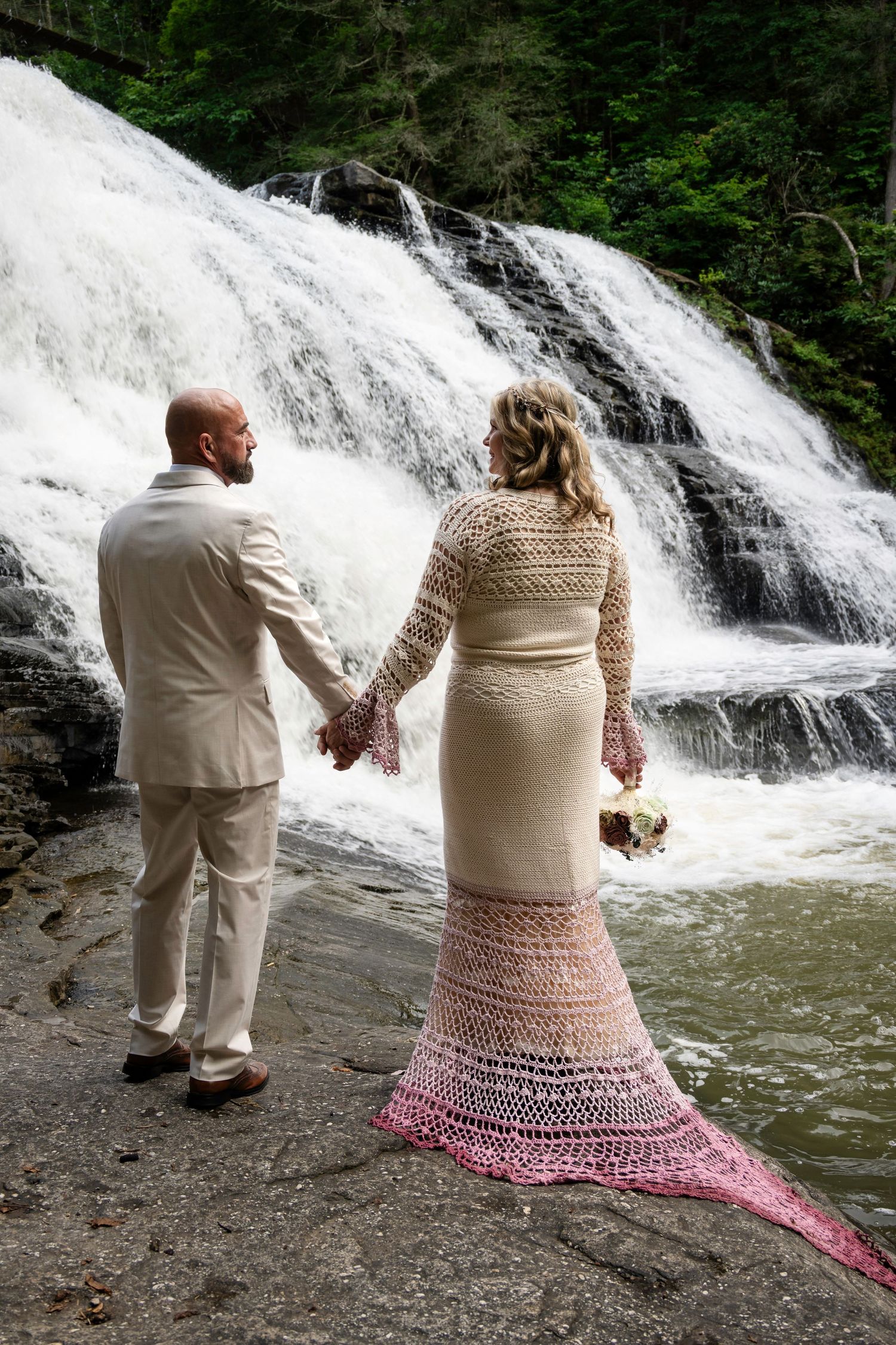 Bride in pink ombre crochet gown and groom in white suit hold hands before a cascading waterfall.