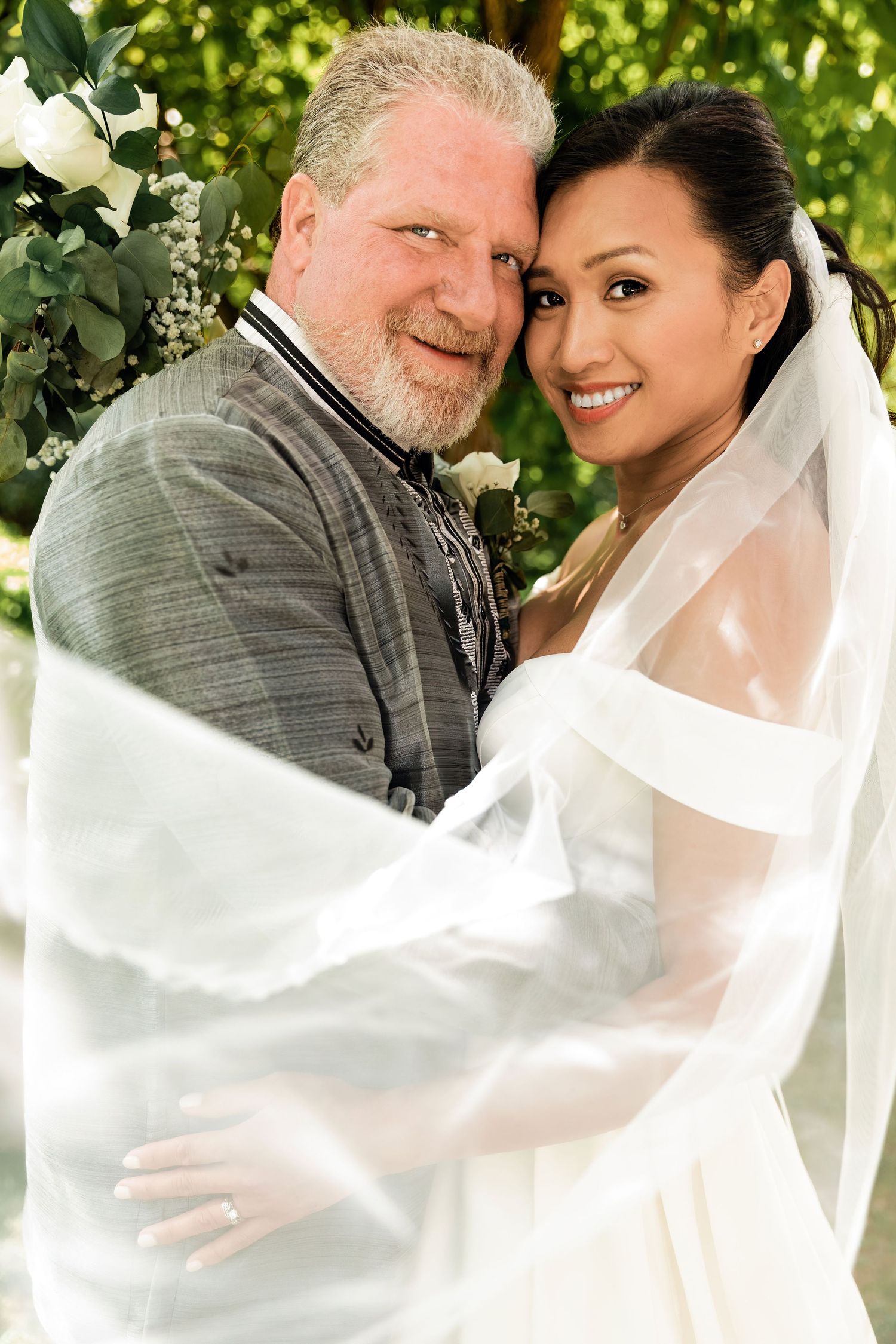 A smiling bride in a white veil and groom in a gray suit embrace outdoors during their wedding ceremony.