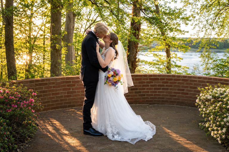 Bride and groom share a kiss on a brick terrace surrounded by trees and a lake at golden hour.