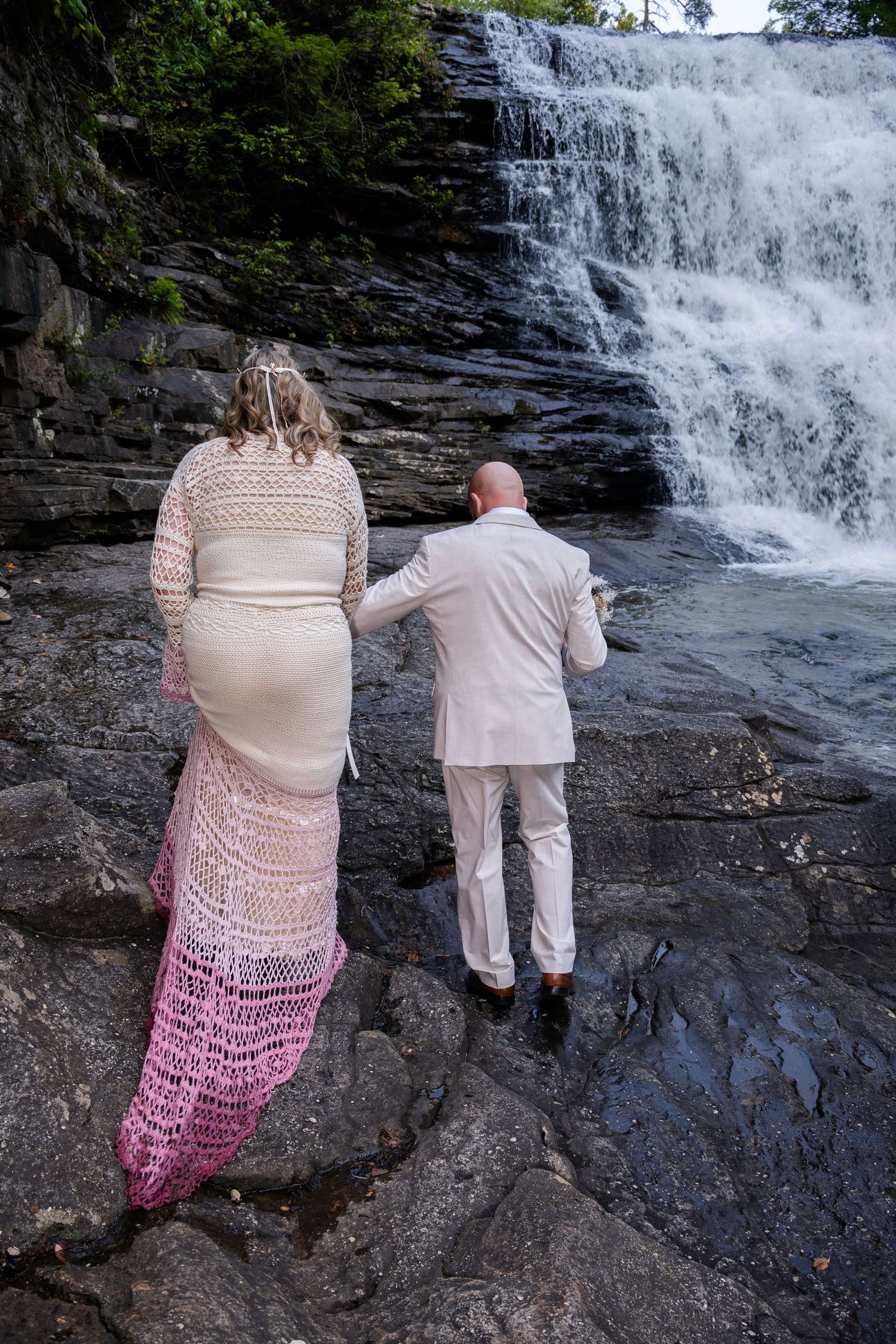 Bride and groom walk hand-in-hand toward a scenic waterfall, bride in crochet dress with pink train, groom in light suit.