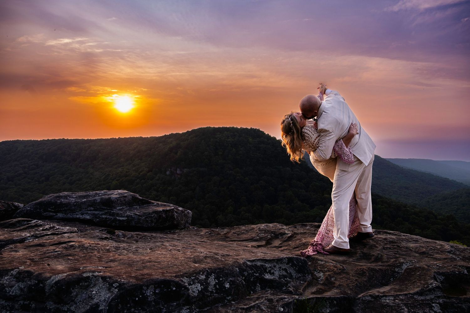 Couple sharing romantic kiss on rocky mountaintop at sunset with colorful sky and forest valley below.