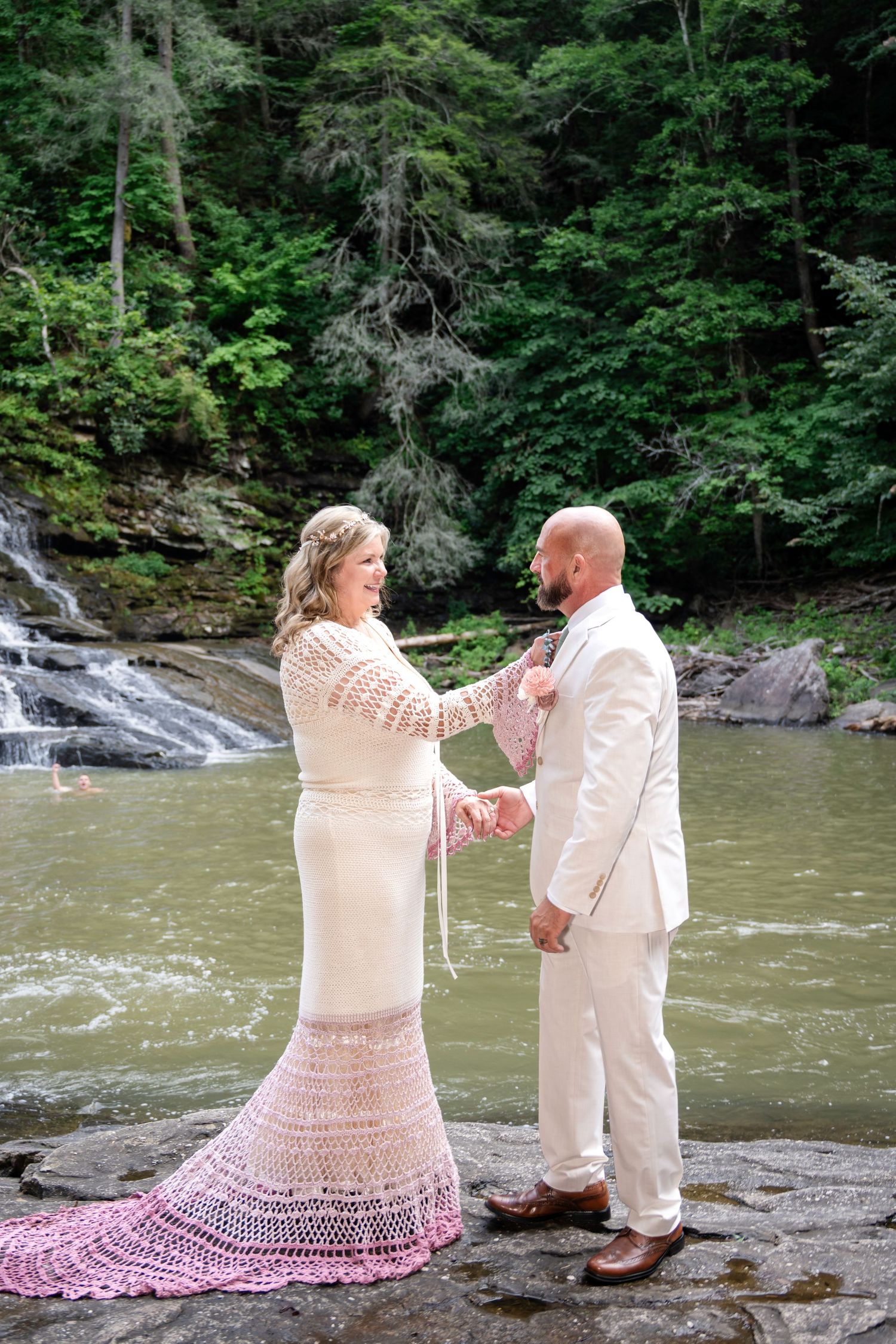 Bride in crochet gown and groom in white suit exchange vows on rocks beside a waterfall in a lush forest.