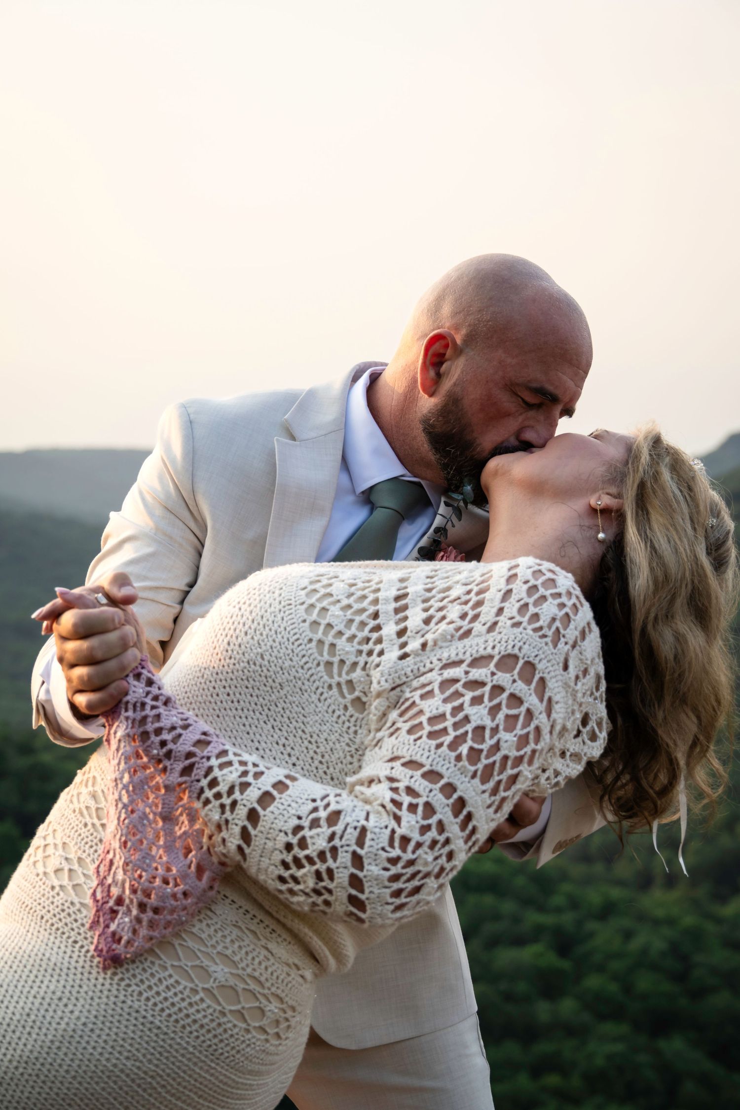 Bride and groom share a romantic dip kiss outdoors at sunset, wearing elegant wedding attire with mountain views.