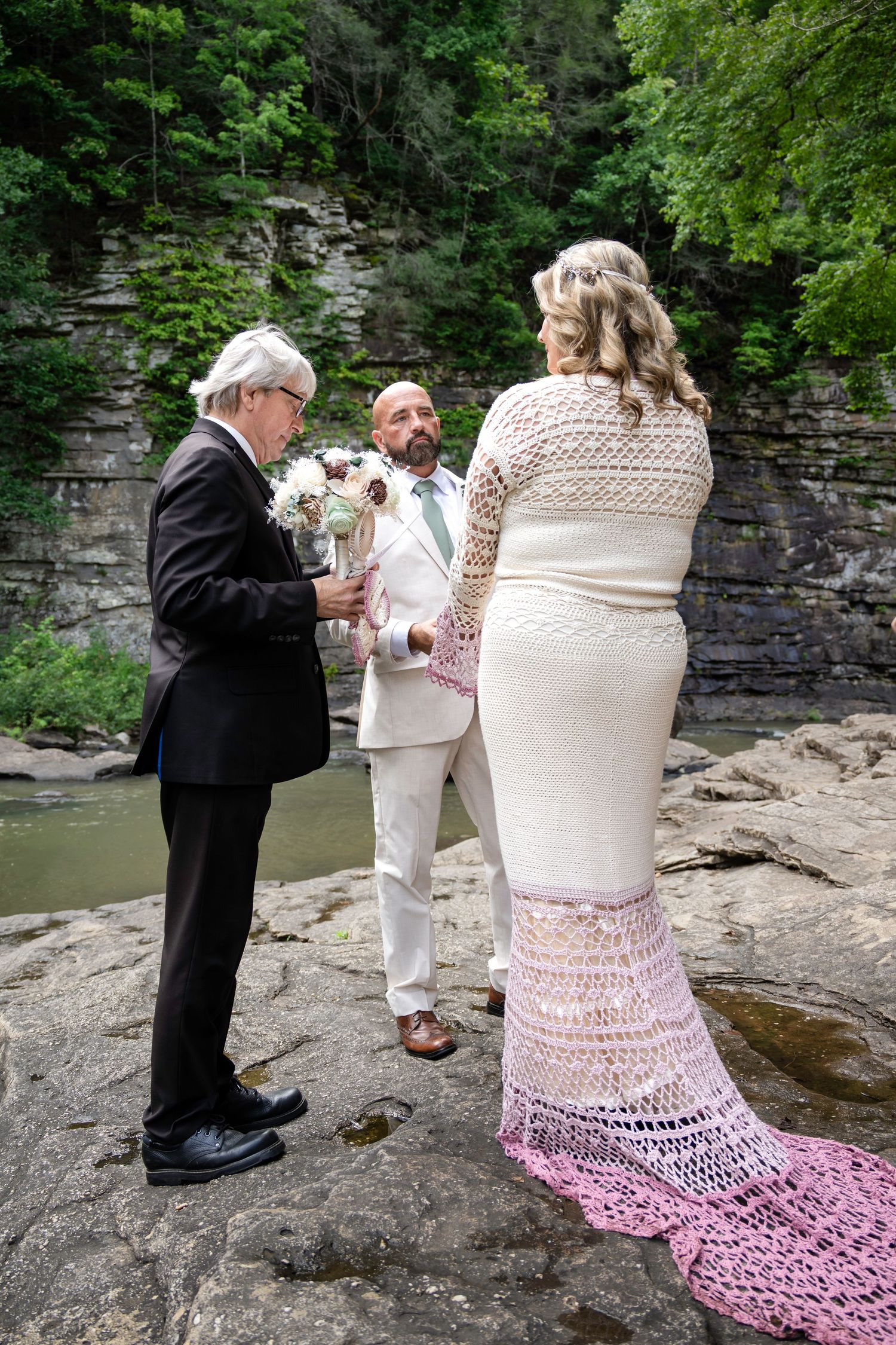 Outdoor wedding ceremony on rocky riverbank with bride, groom, and officiant surrounded by lush green cliffs.