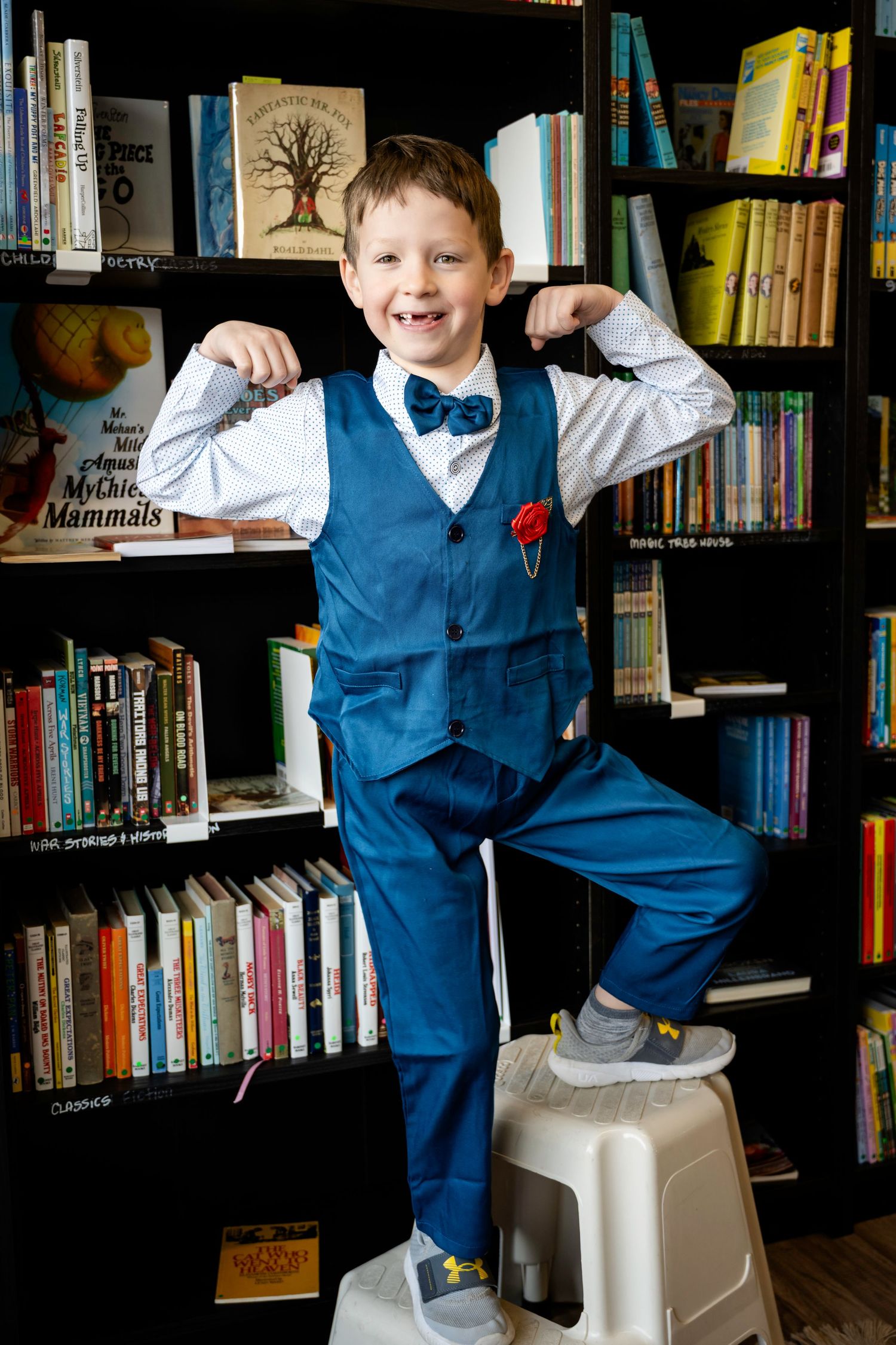 Young boy in blue suit flexing muscles on stool in front of colorful bookshelf library.